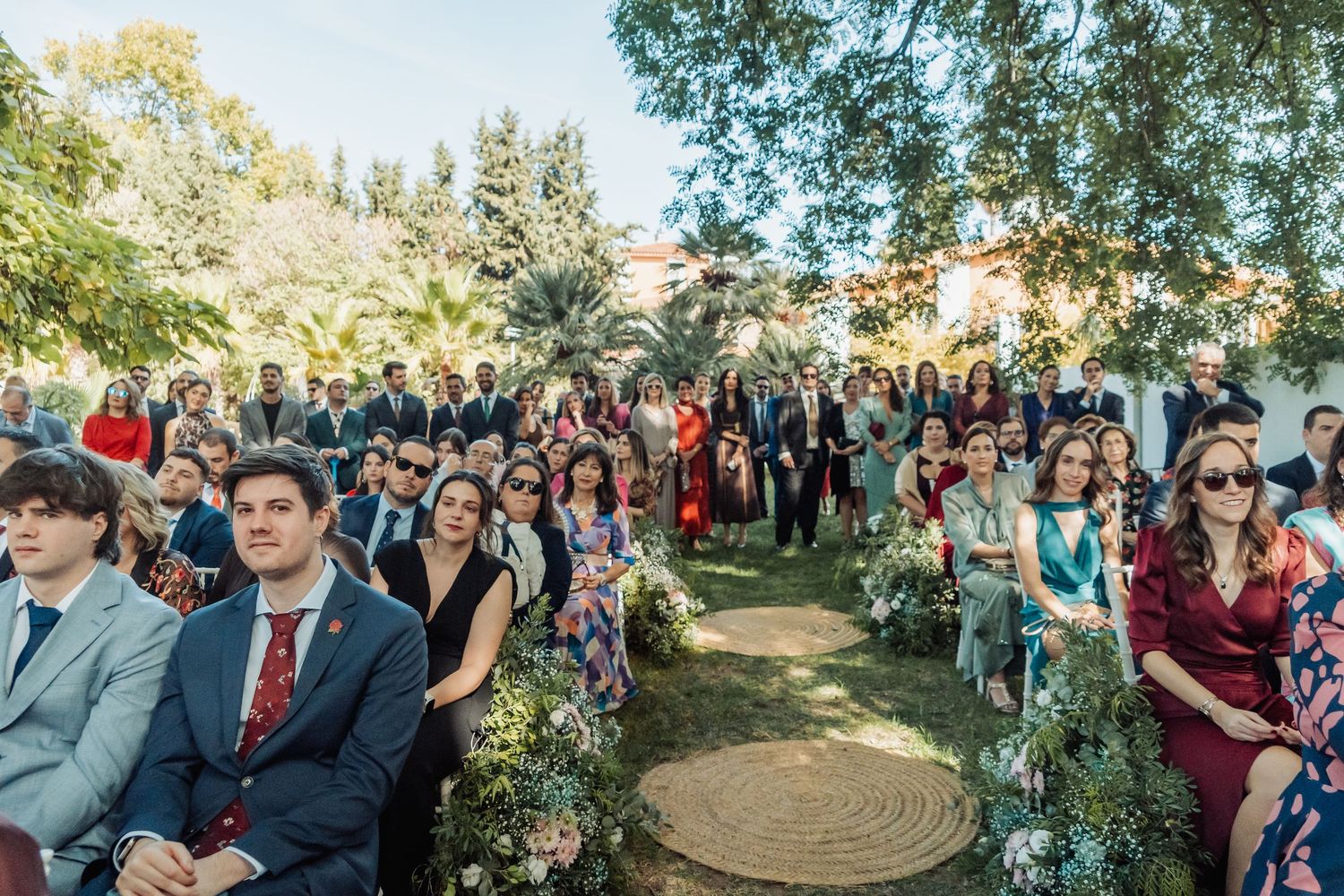 Large group of elegantly dressed wedding guests seated in white chairs at an outdoor garden ceremony.