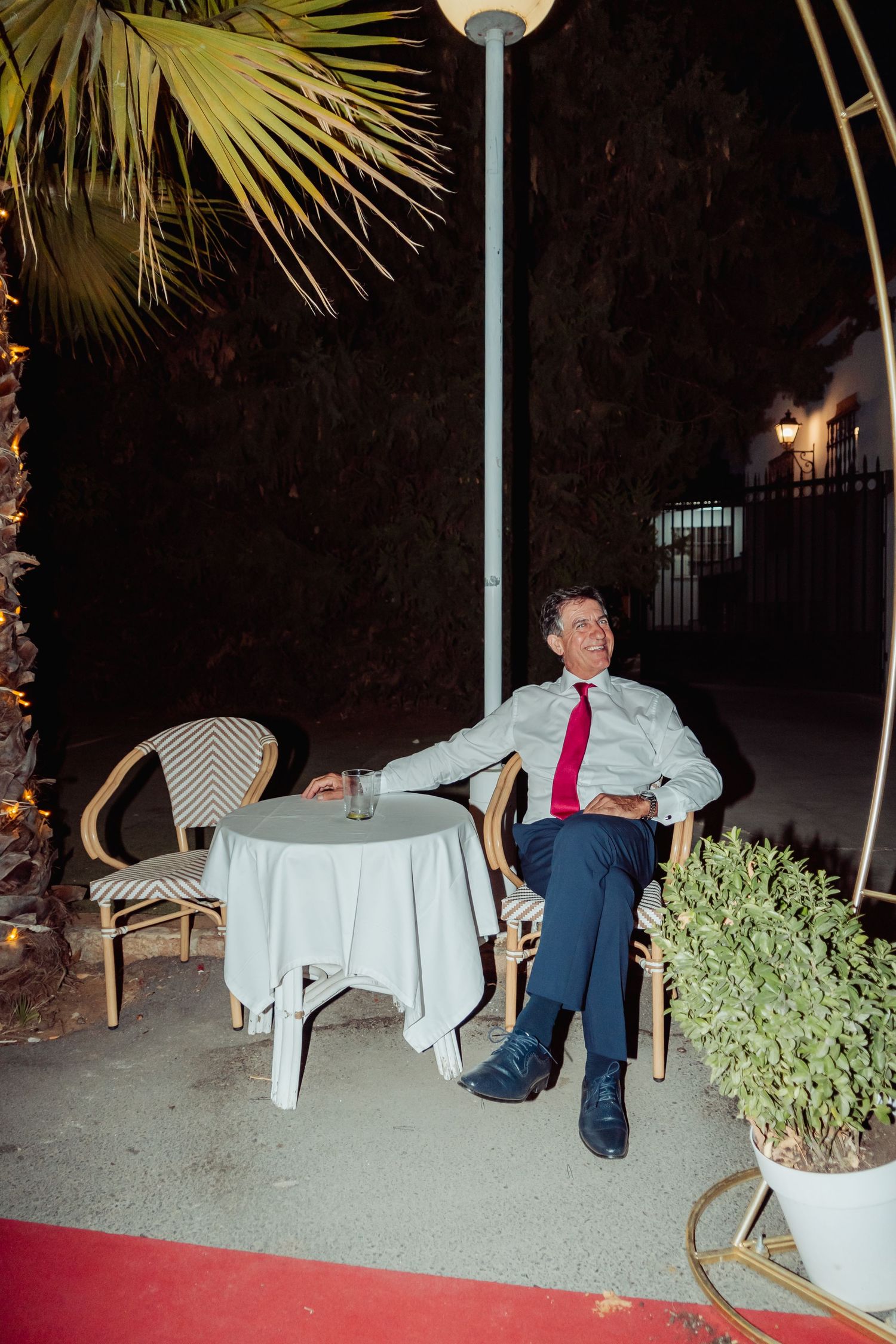 Man in white shirt and red tie relaxing alone at an outdoor table near a palm tree at night.