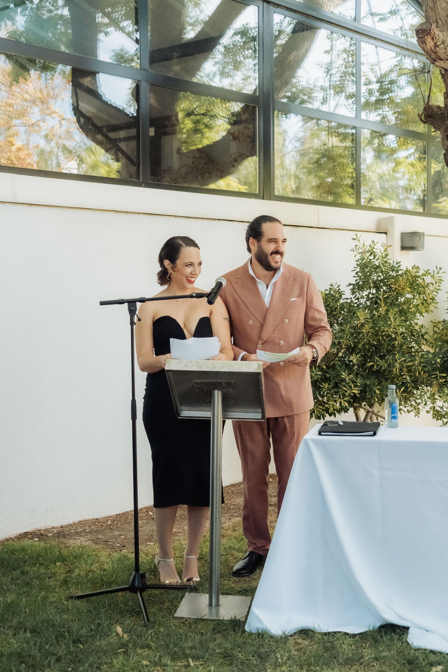 A smiling couple in formal attire stands together at a podium delivering a speech at an outdoor wedding.