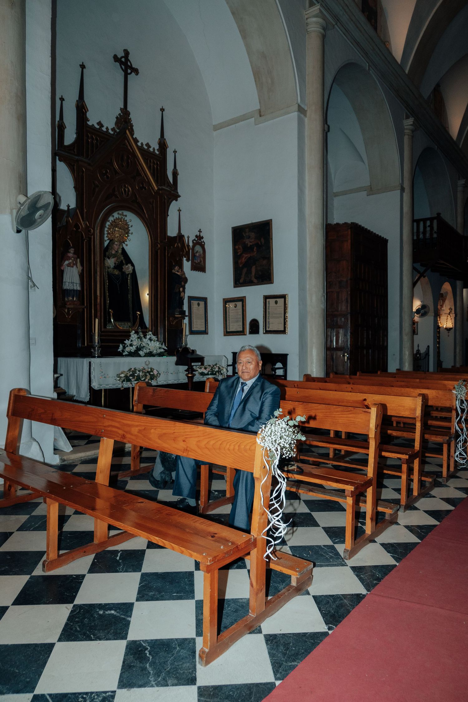 Elderly man in suit sits alone on wooden pew in a historic church decorated for a wedding.