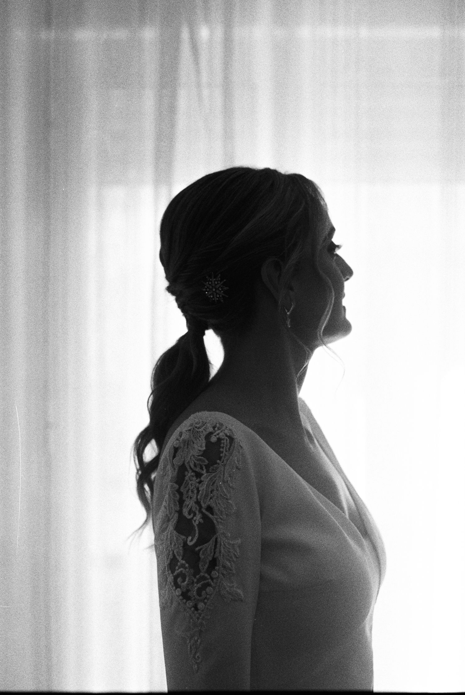 Black and white silhouette of bride with braided hair and lace sleeve dress by window.