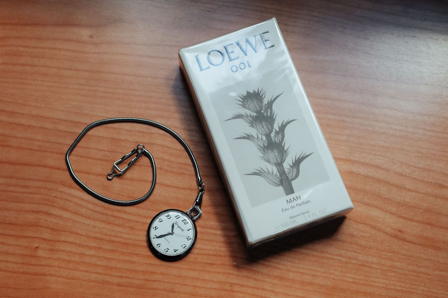 Loewe 001 fragrance box beside vintage pocket watch on wooden surface.