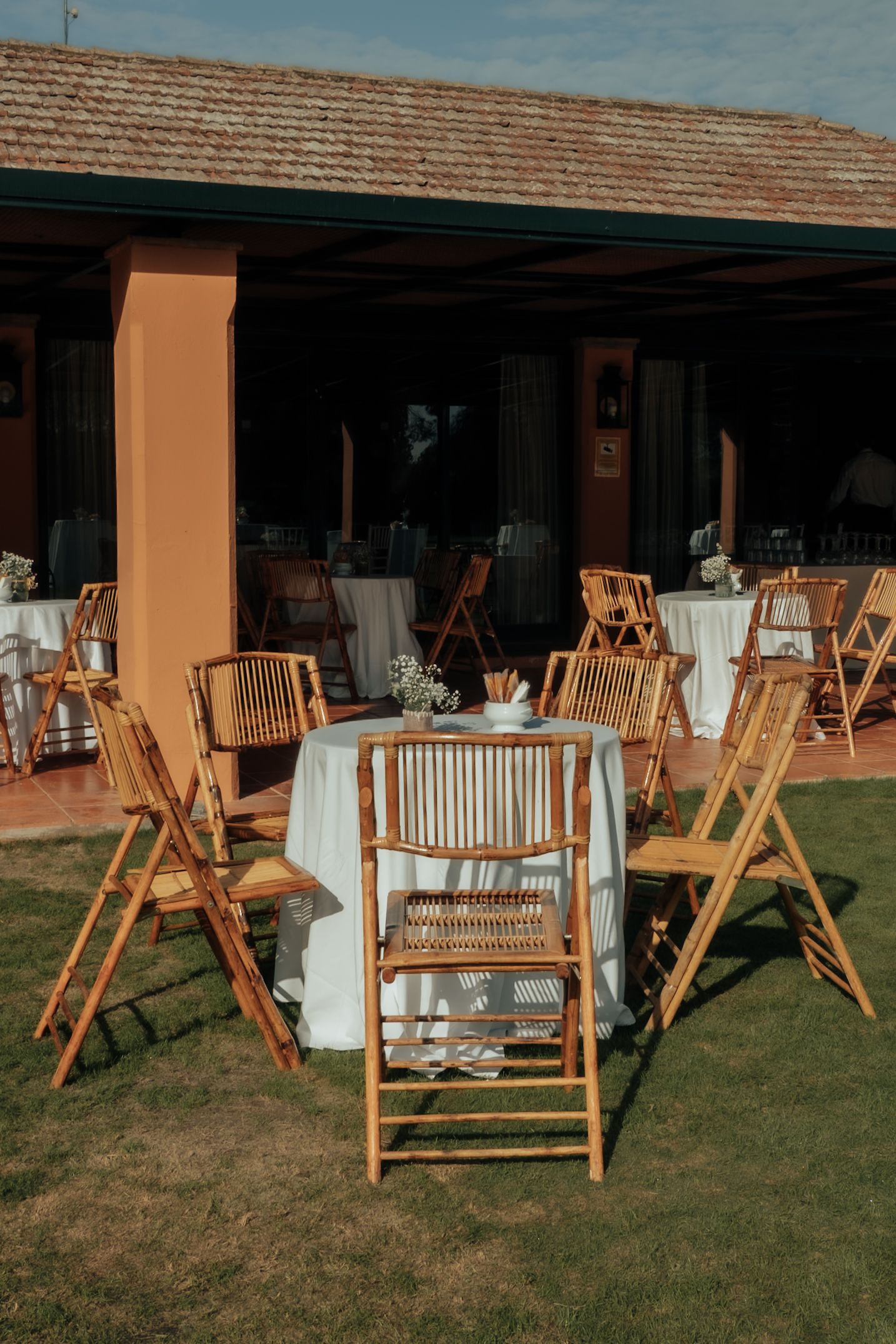 Outdoor wedding reception setup with bamboo chairs and white tablecloths on green lawn near terracotta building.