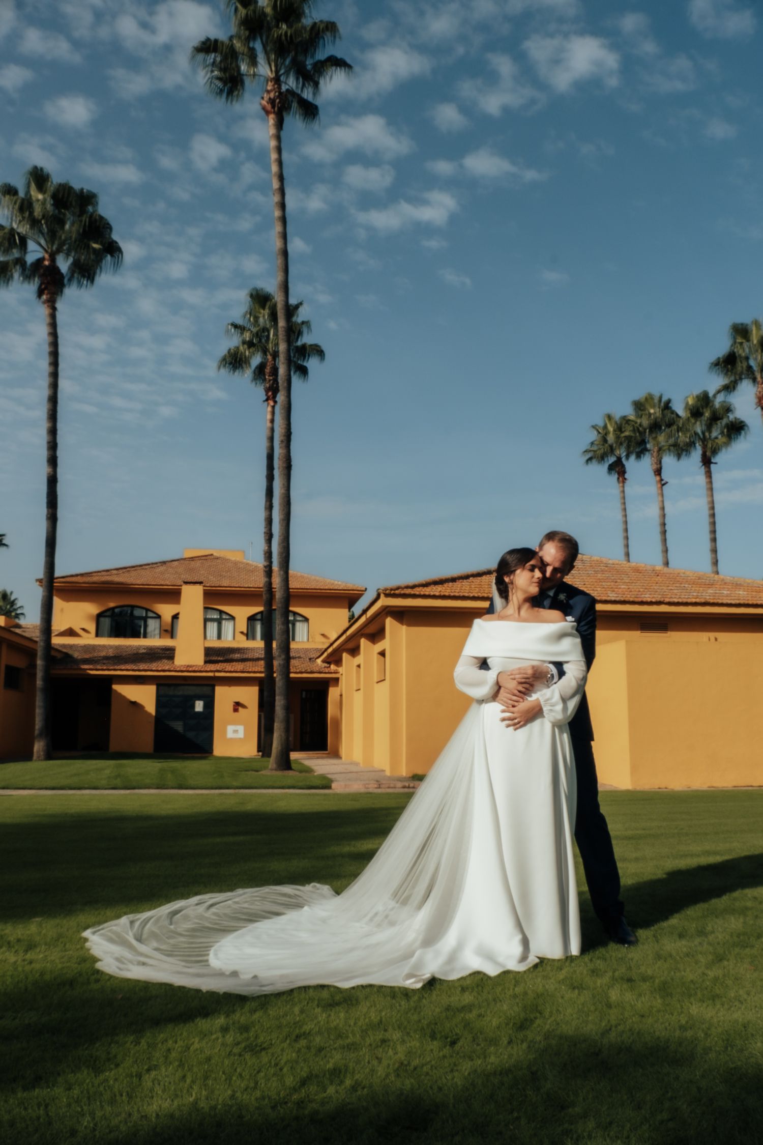 Bride in off-shoulder white gown with long train embraced by groom in navy suit near palm trees.