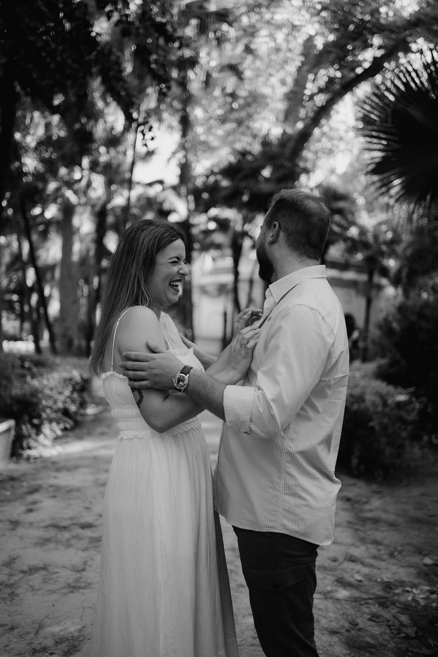 Black and white photo of laughing bride in white gown embracing bearded groom among garden trees.