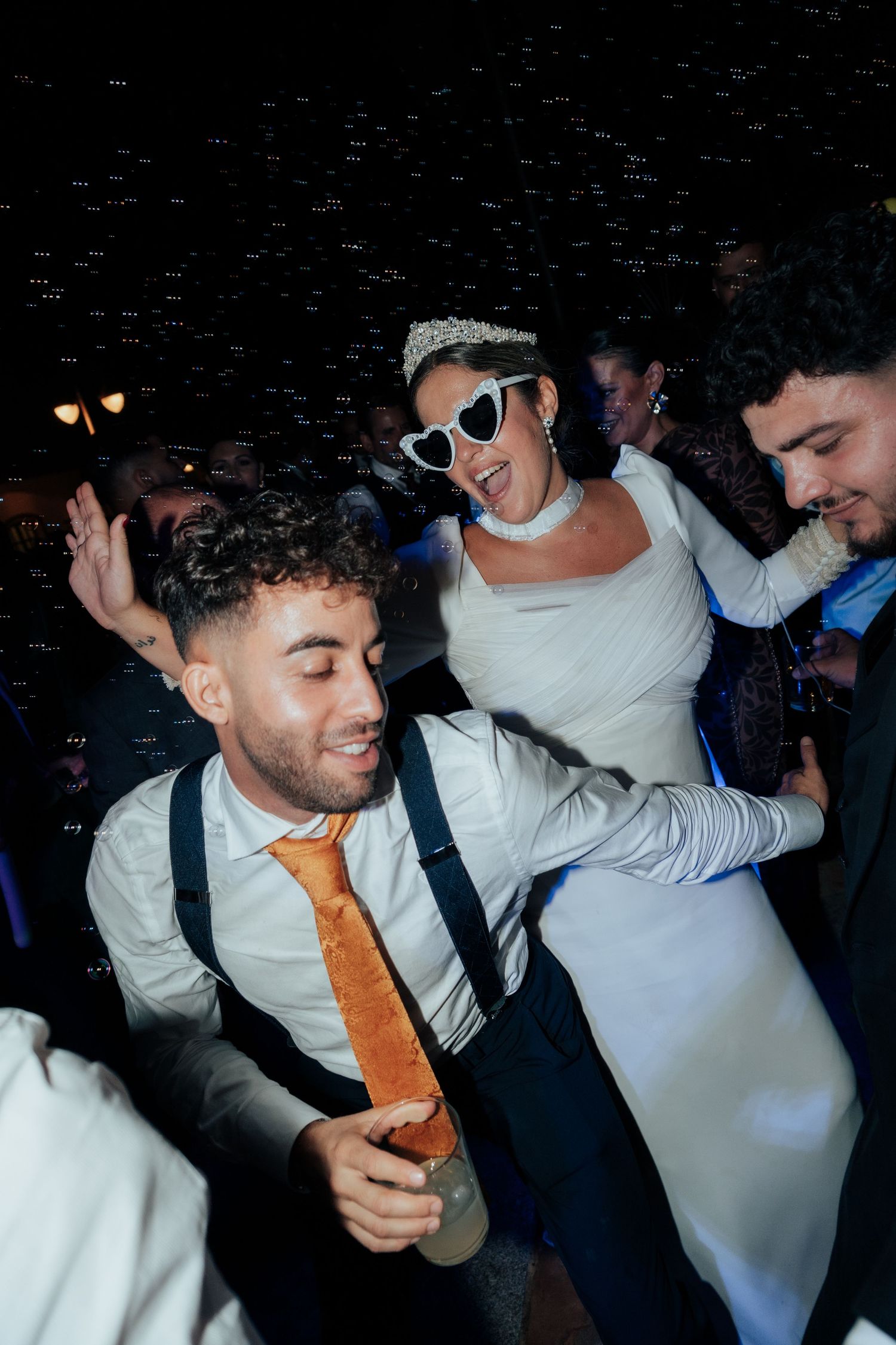 Bride in white dress and heart sunglasses dances joyfully with guests at a lively wedding party.