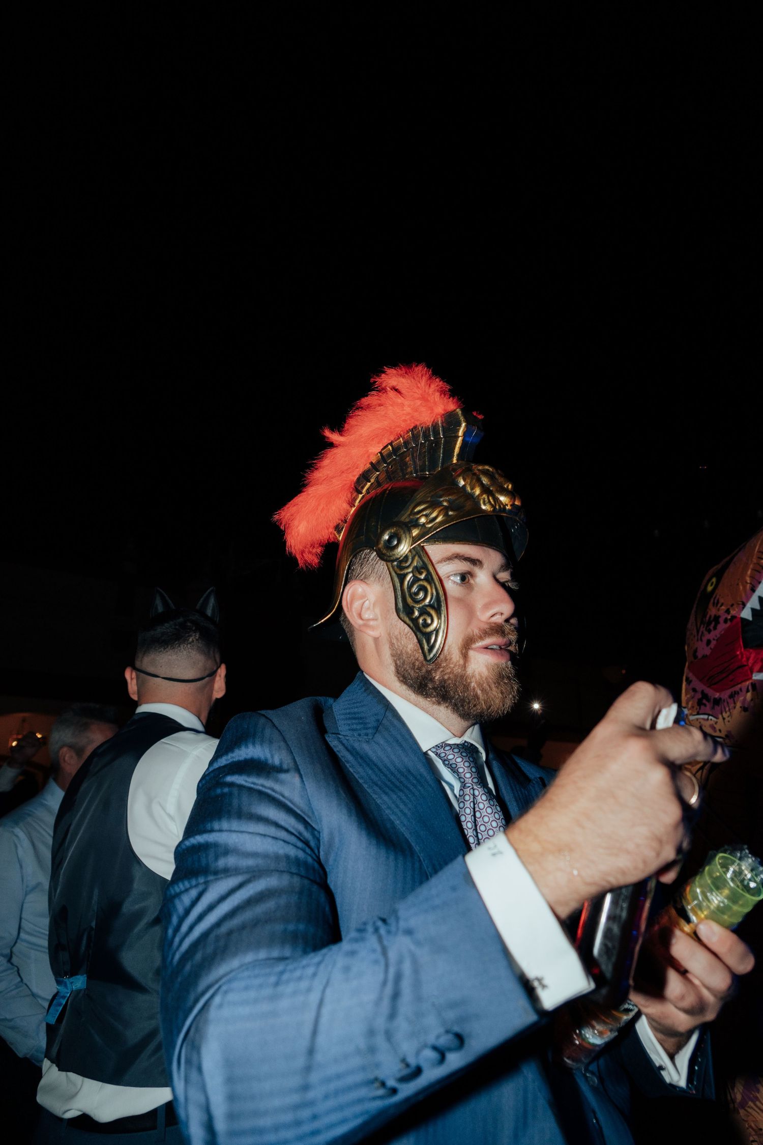 Man in a blue suit wearing a decorative Roman gladiator helmet at a festive nighttime wedding party.