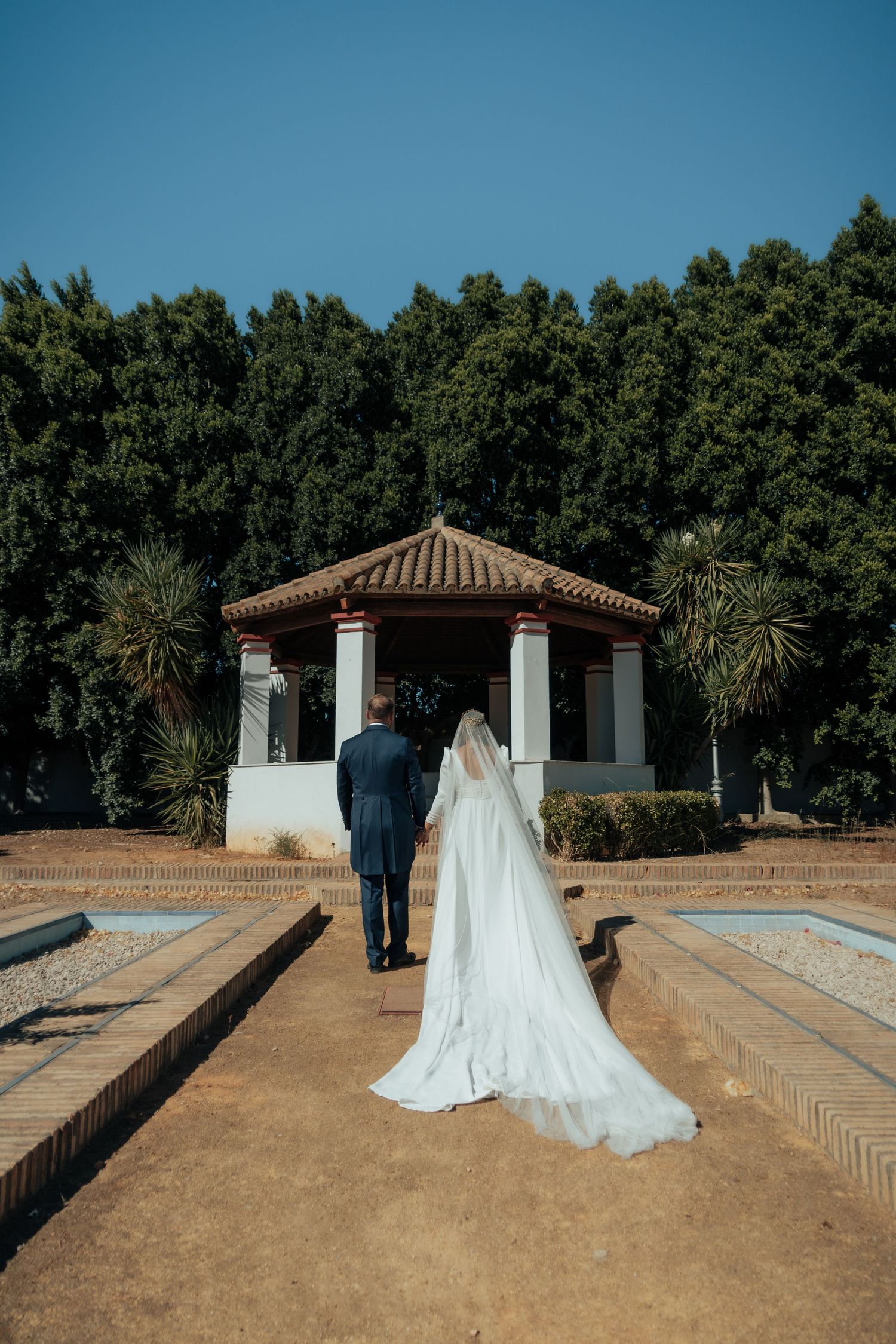 Bride and groom walking hand in hand toward a red-roofed gazebo surrounded by tall green trees.