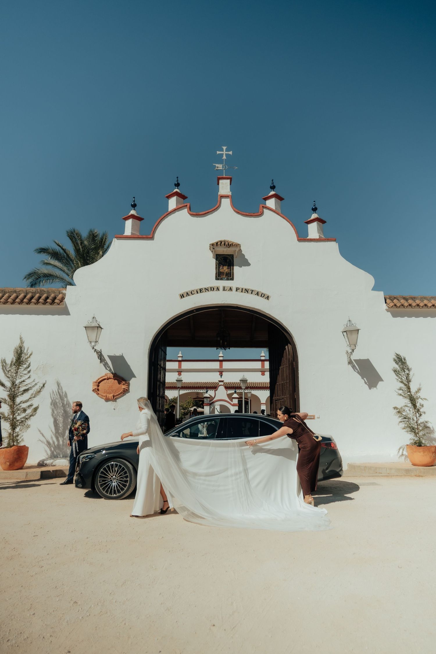 Bride arrives at Hacienda La Pintada as assistant arranges her long white veil by luxury car.