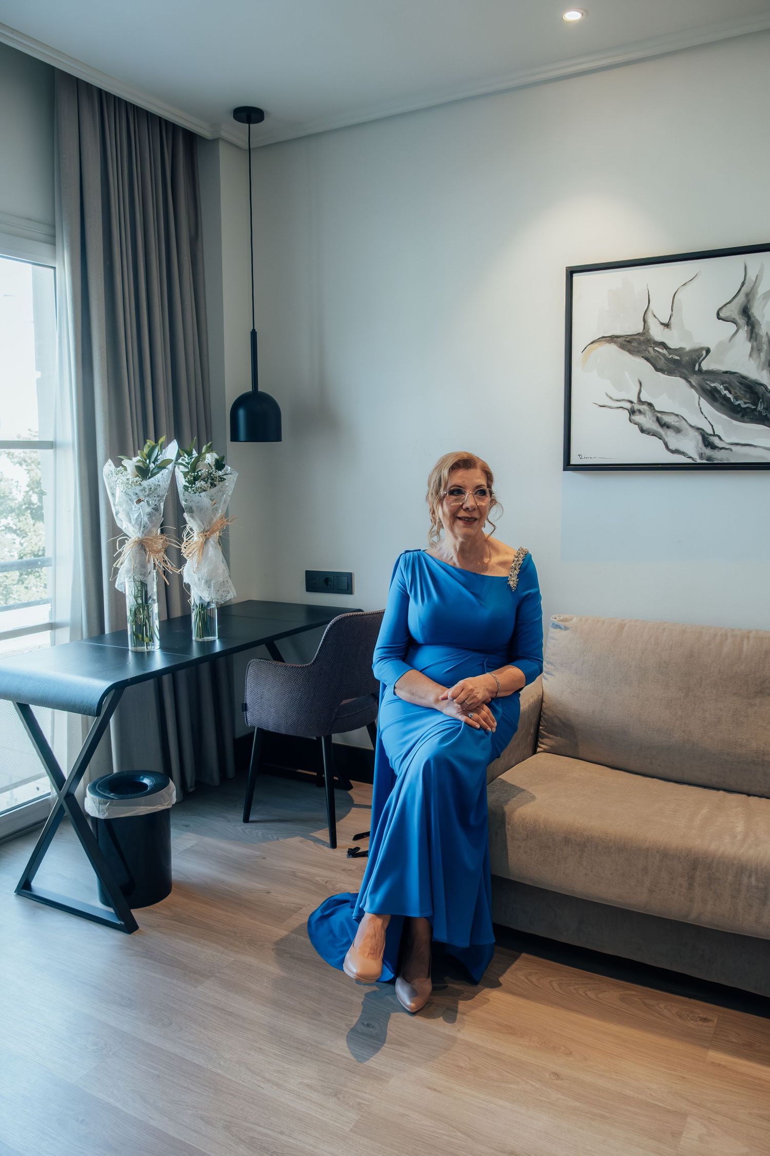 Elegant older woman in a royal blue gown sits gracefully on a sofa in a modern hotel room.