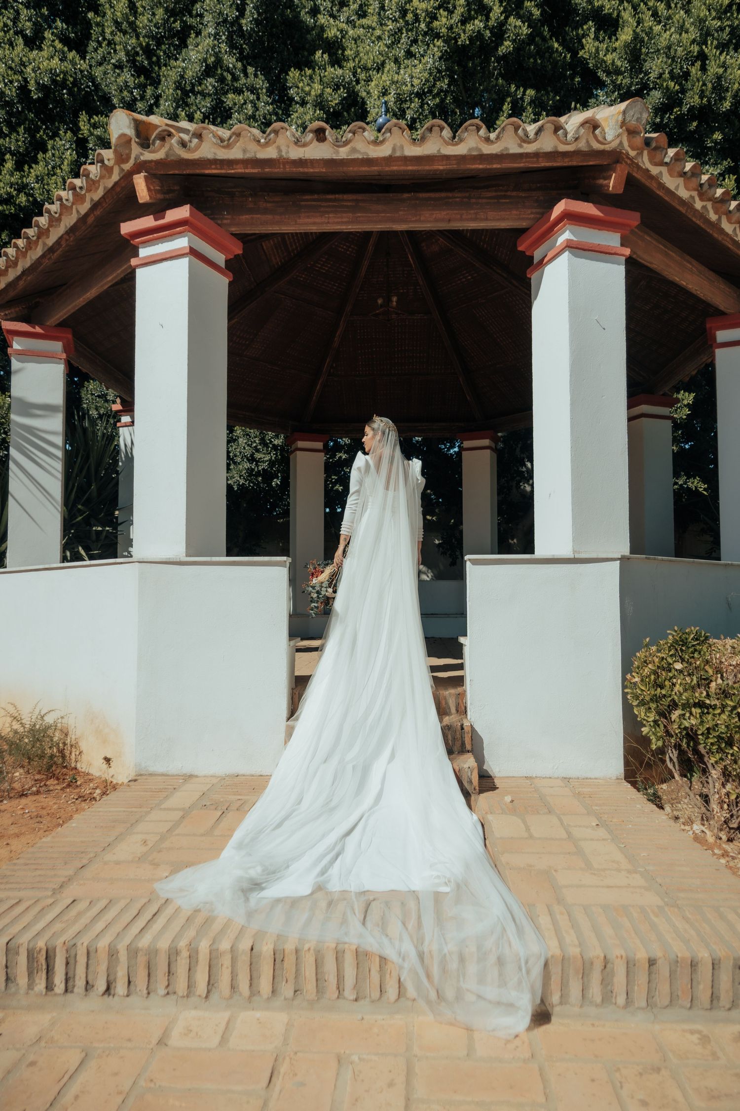 Bride in elegant white gown with long cathedral veil stands before a Spanish-style gazebo with red columns.
