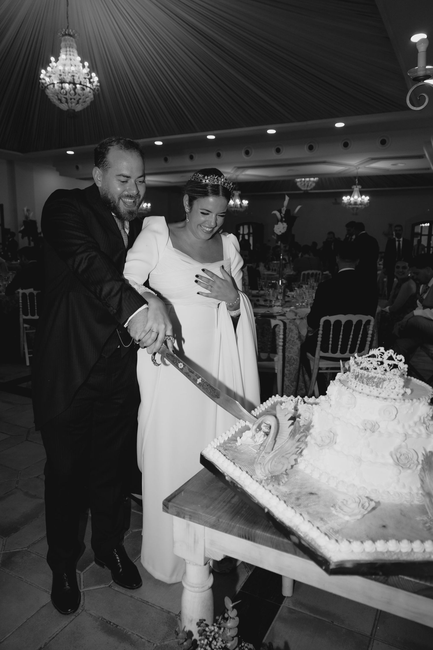 Bride and groom cutting elaborate tiered wedding cake together at reception in black and white.