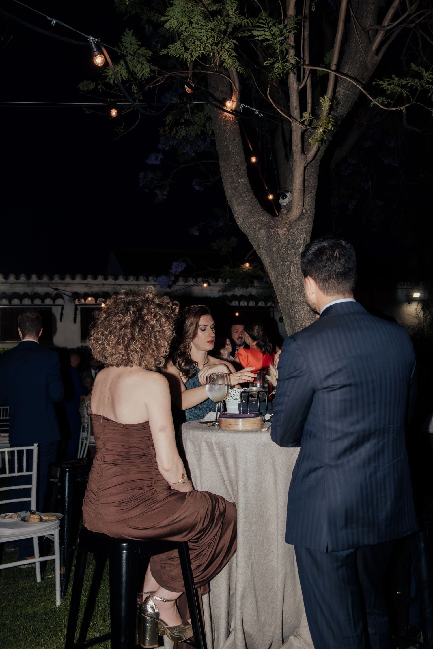 Guests socializing at a candlelit outdoor evening reception under string lights and trees.