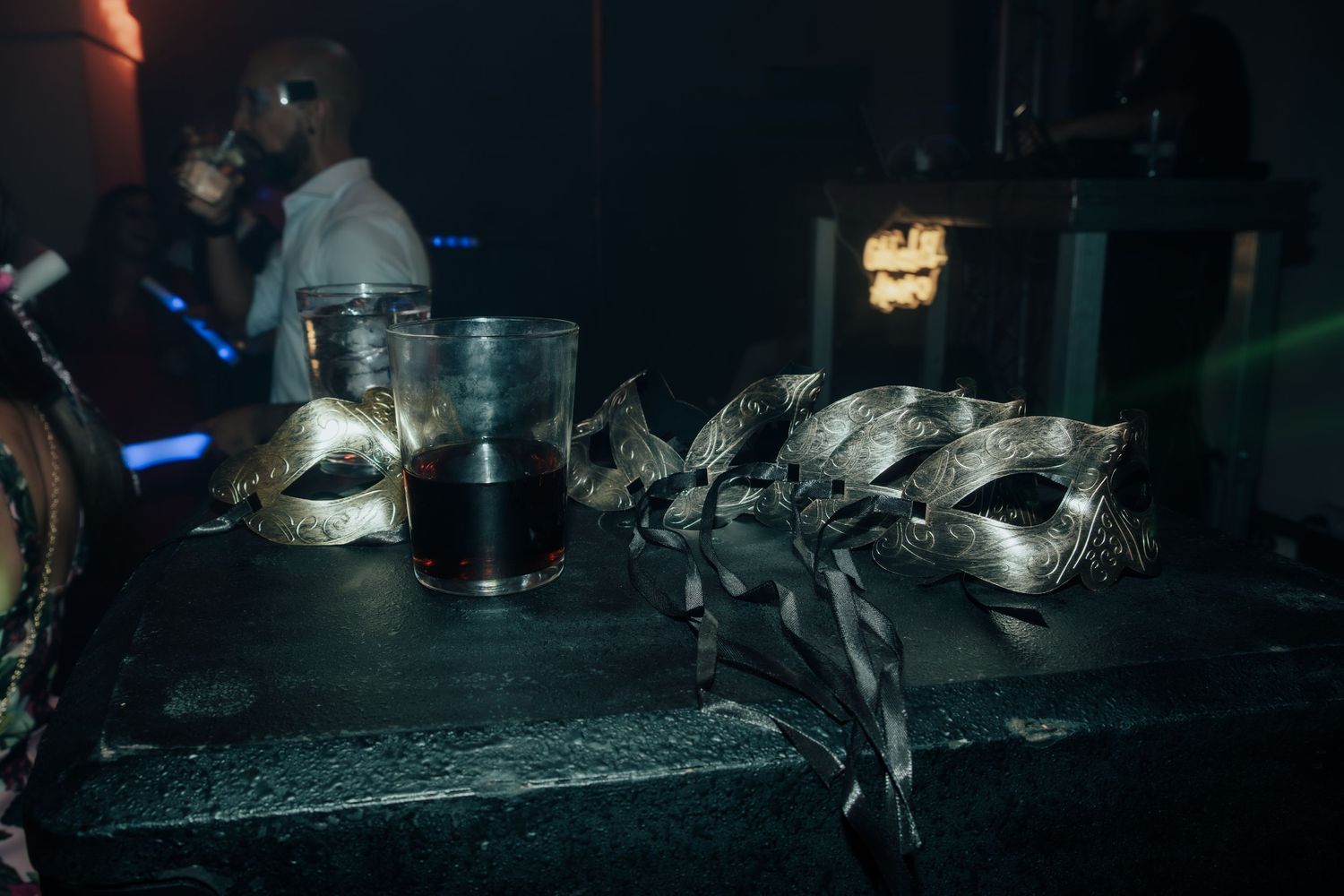 Two ornate masquerade masks rest on a dark surface beside a drink glass at a party.
