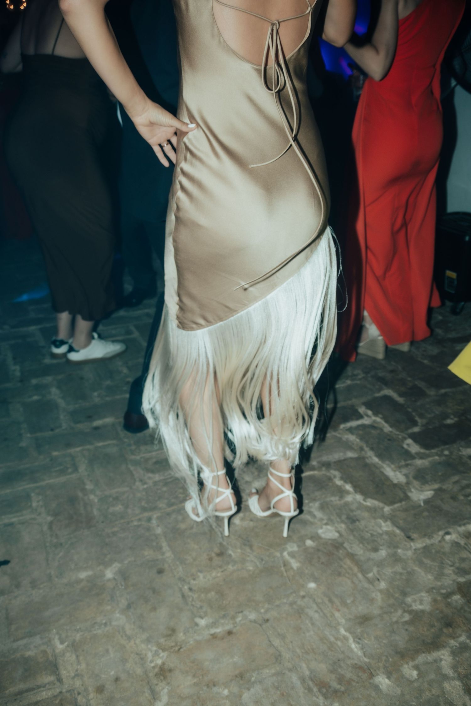 Woman in elegant gold fringe dress with white heeled sandals dancing on cobblestone floor at party.