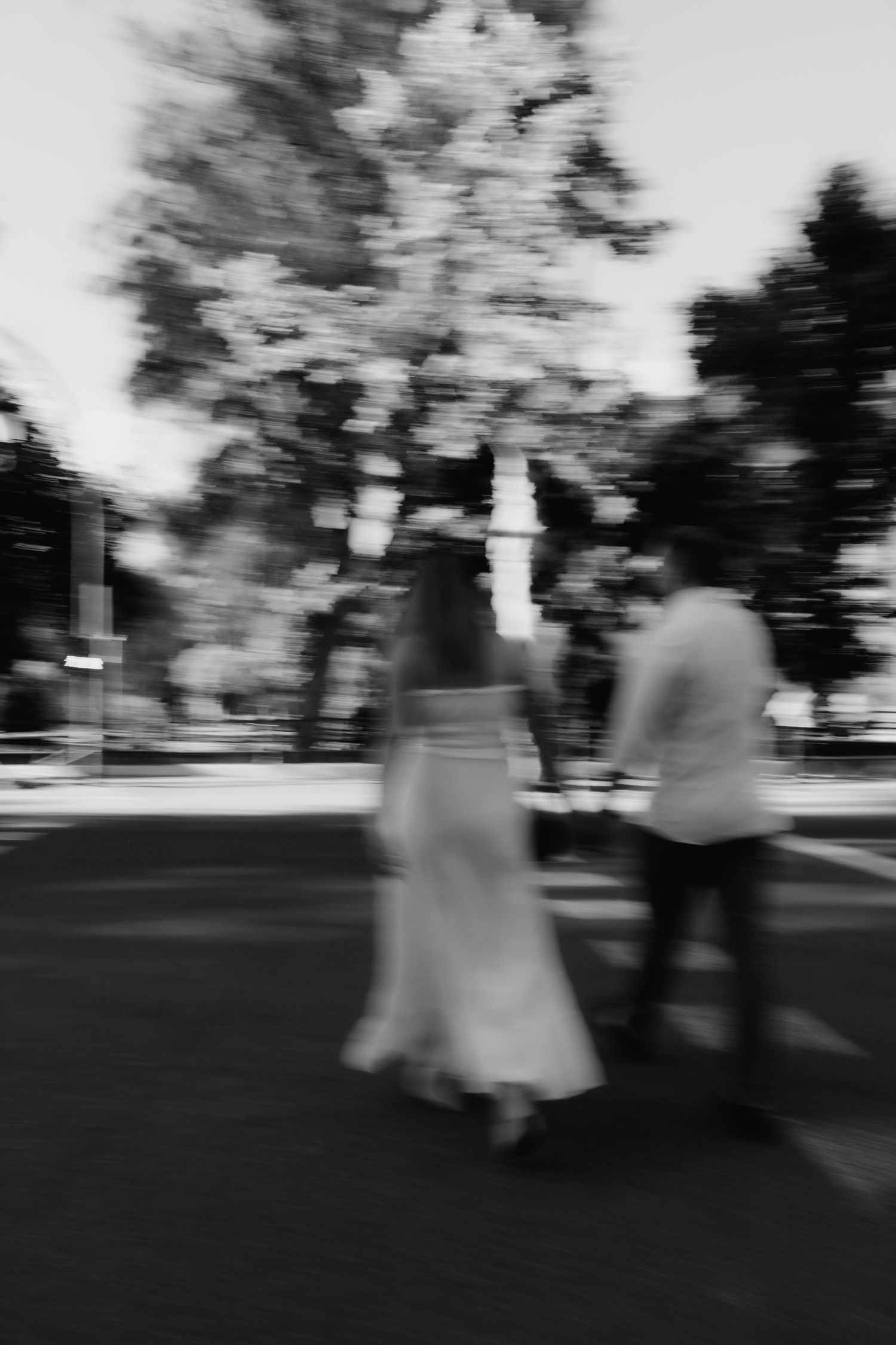 Artistic blurred black and white photo of bride and groom walking hand in hand through a tree-lined path.
