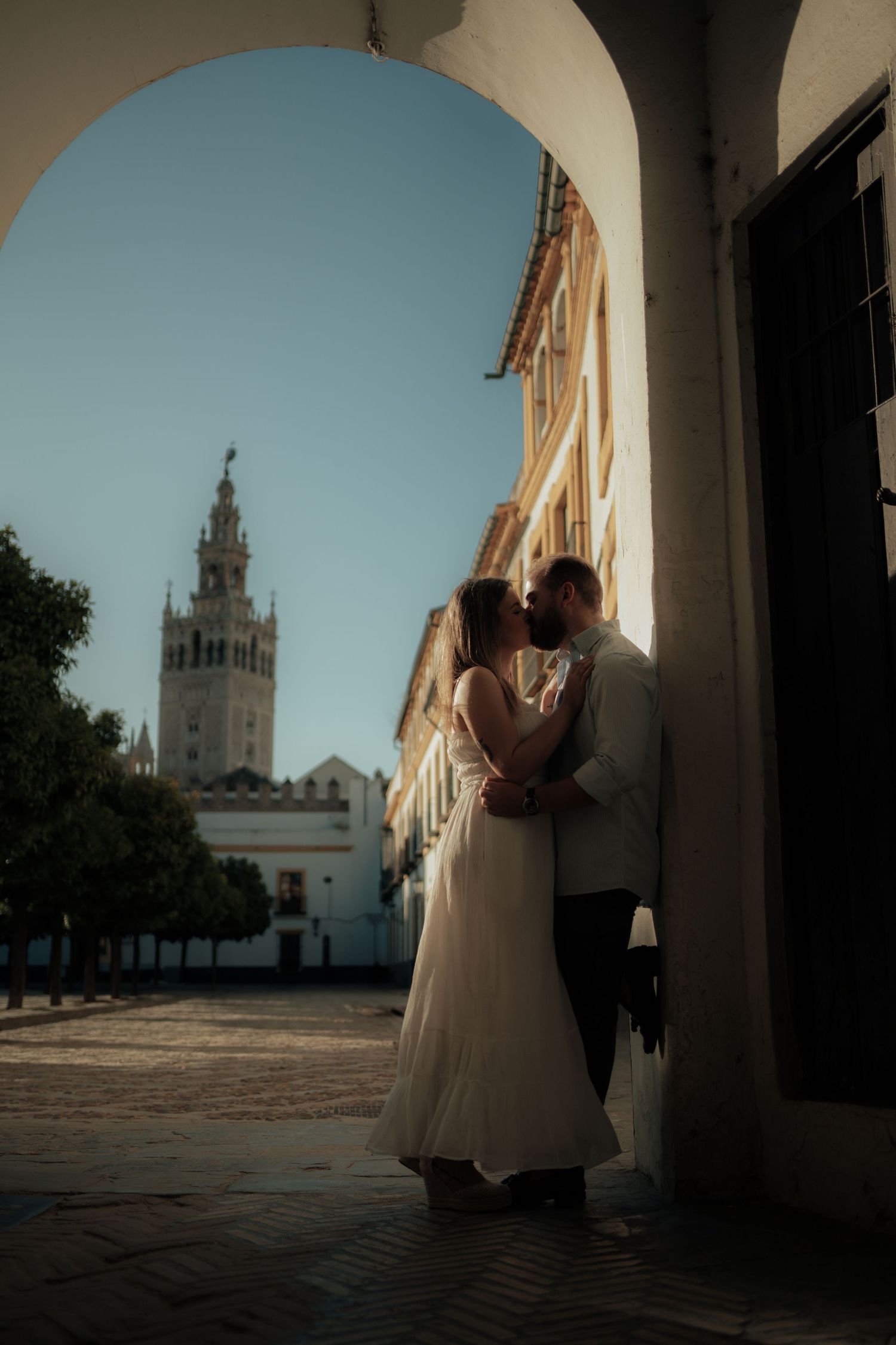 Bride and groom sharing a romantic kiss under an arch with the Giralda tower of Seville in background.