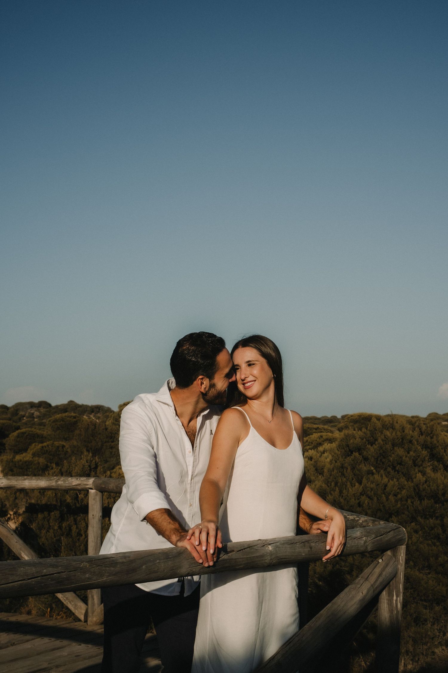 Romantic couple sharing a tender moment at a wooden railing overlook at sunset, woman in white dress smiling joyfully.
