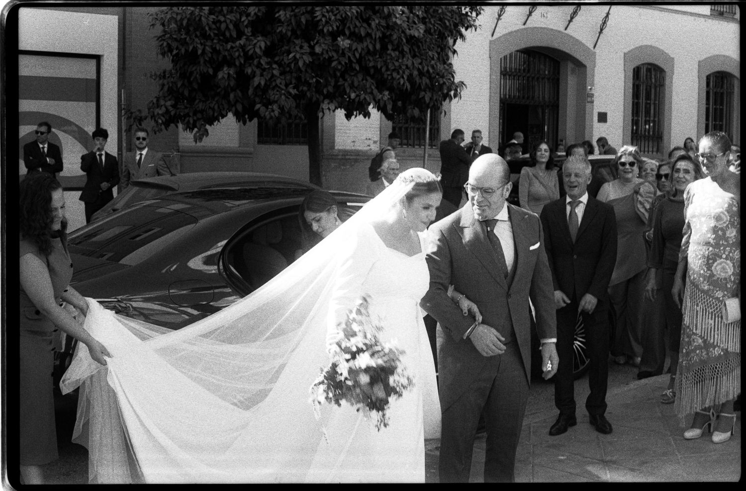 Black and white photo of bride in lace gown with long veil walking with escort outside church.