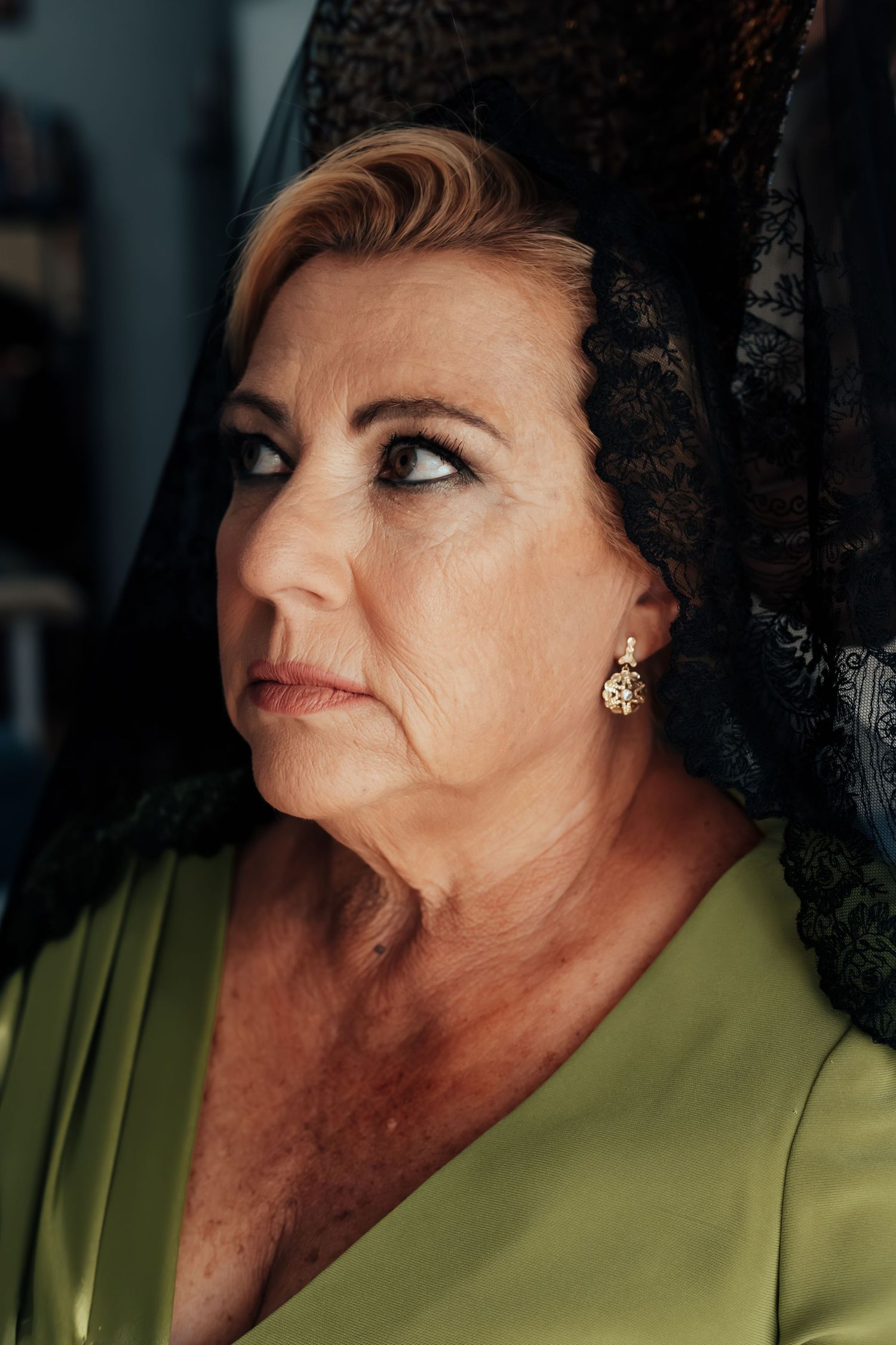 Elegant older woman wearing black lace mantilla veil and green dress with gold earrings.