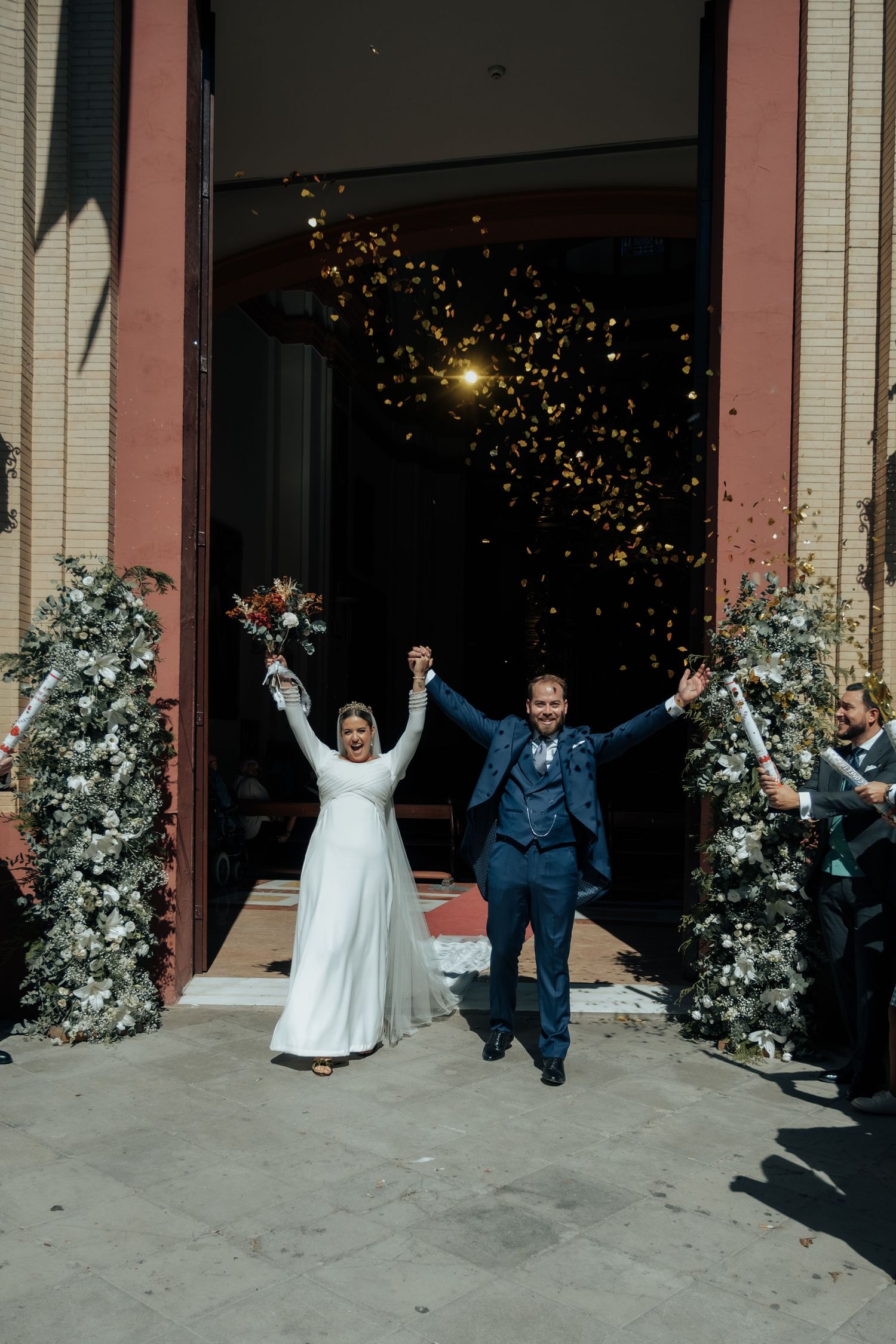 Joyful newlyweds raise hands as confetti showers them exiting church after wedding ceremony.