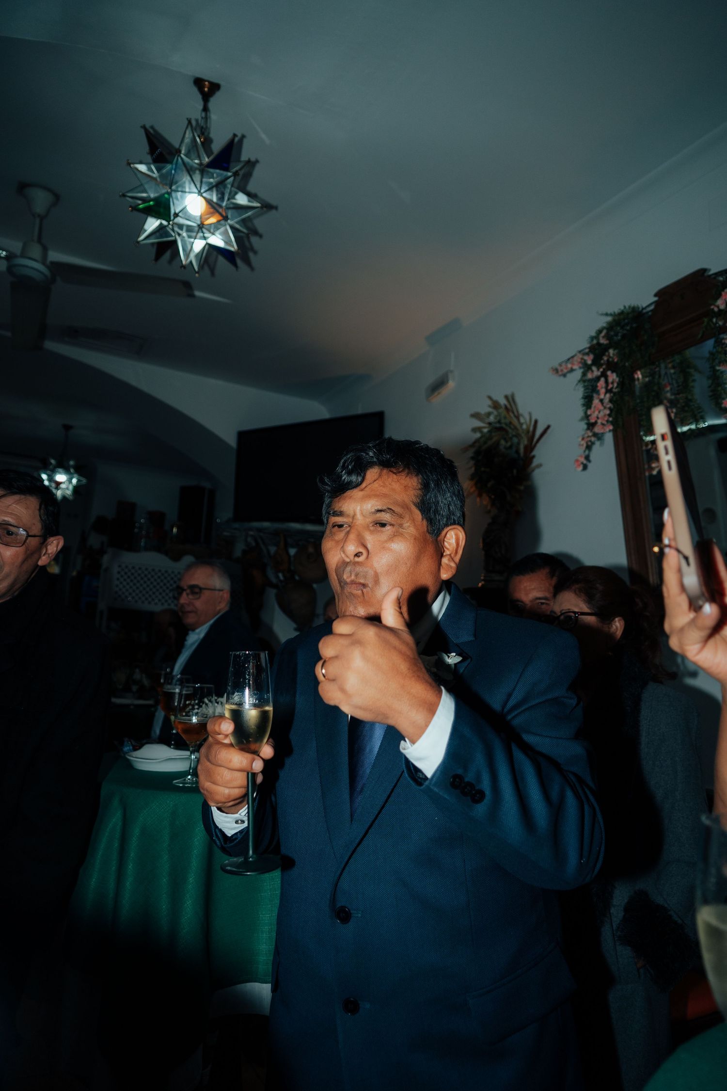 Man in navy suit holding champagne glass and giving thumbs up at an indoor celebration.