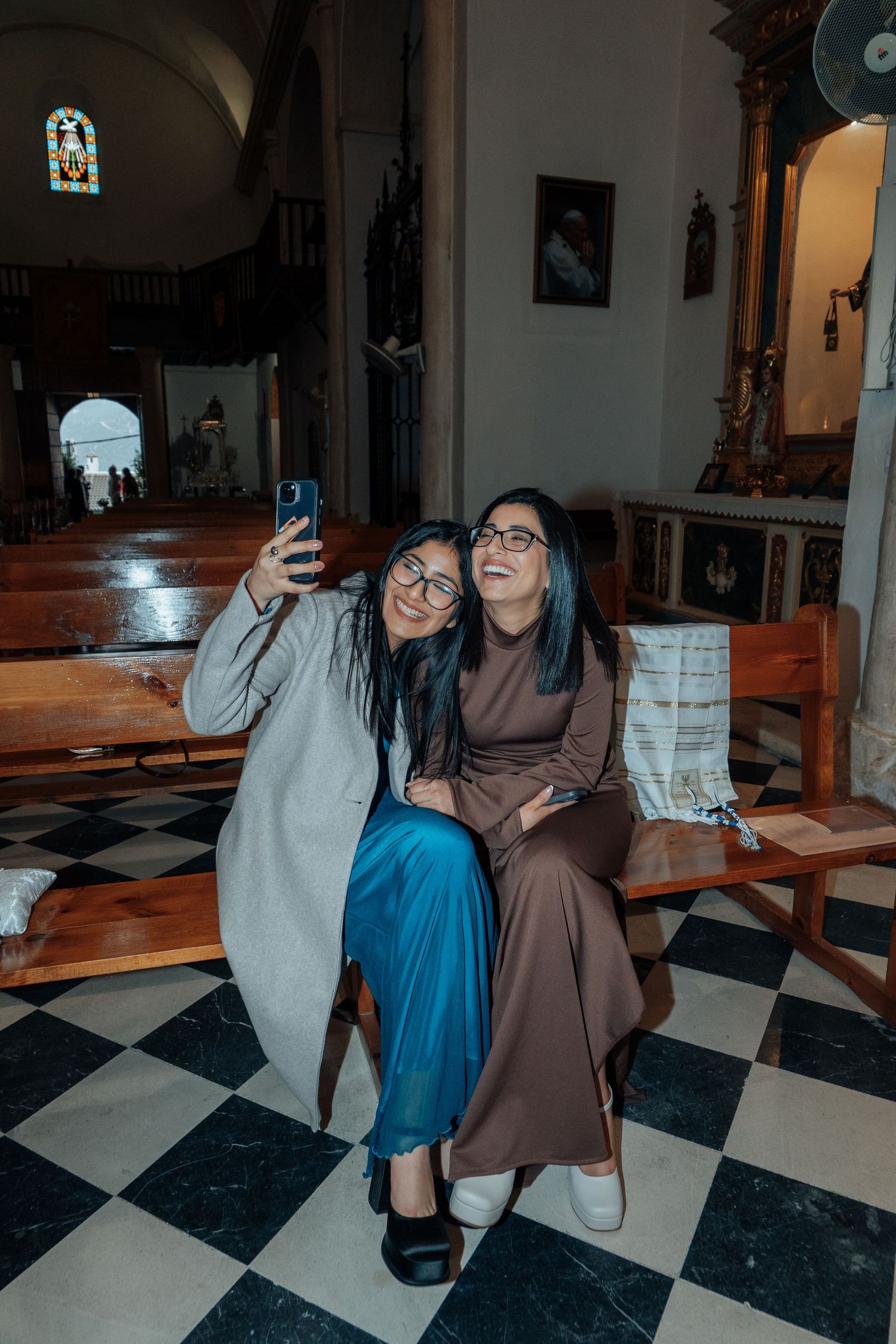 Two women wearing glasses take a selfie together while seated on a church pew during a wedding.