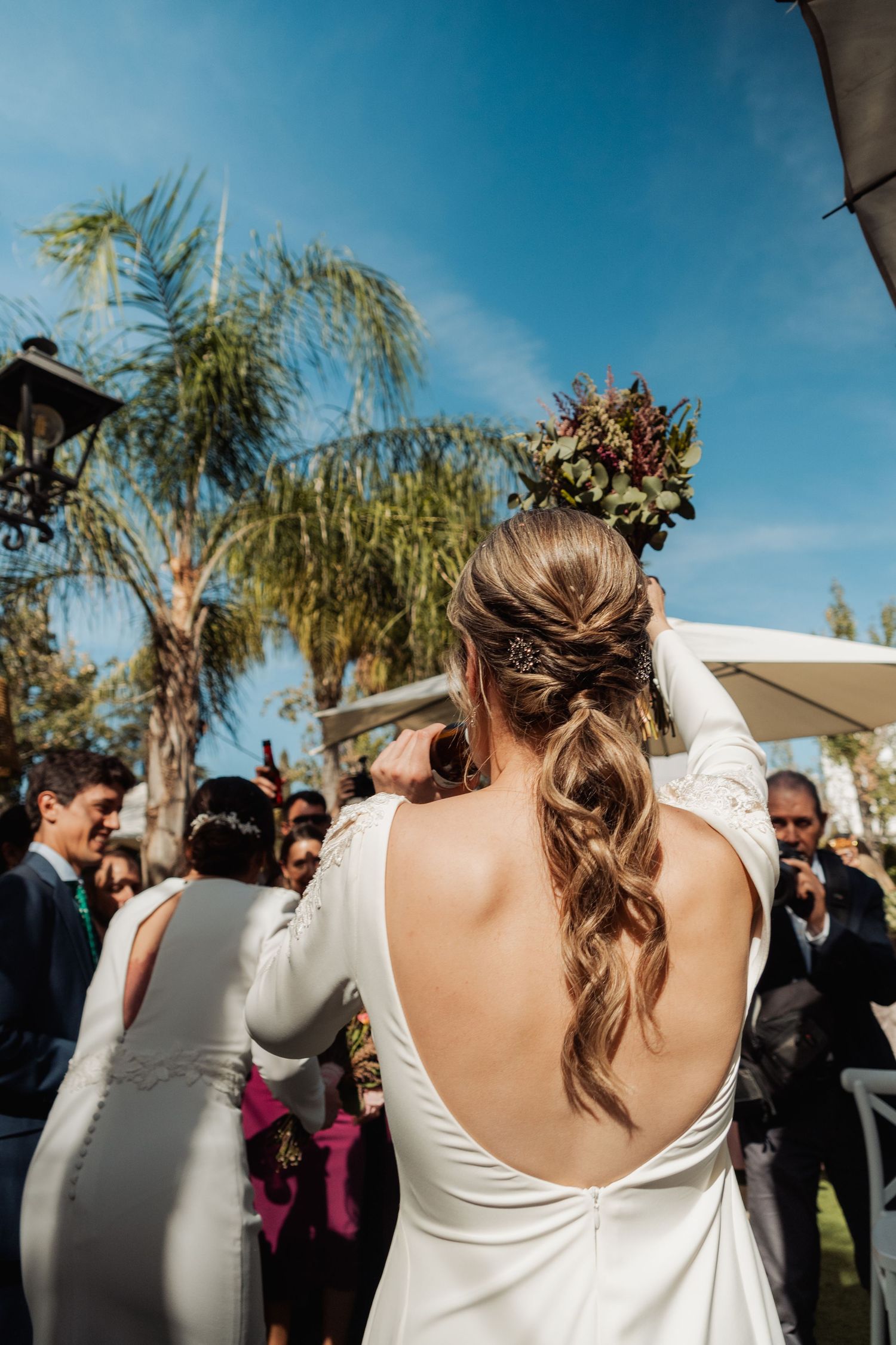 Bride in backless white gown raises bouquet at outdoor wedding celebration under blue sky.