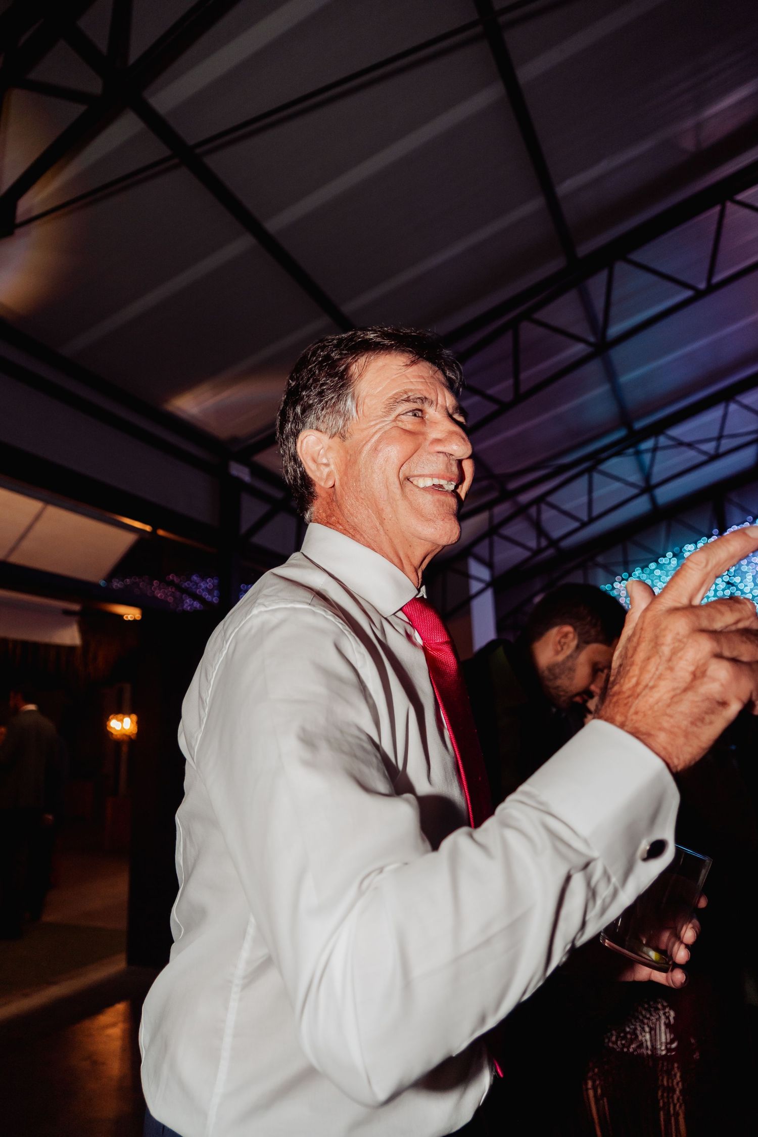 Smiling older man in white suit and red tie enjoying a celebration at an indoor evening event.
