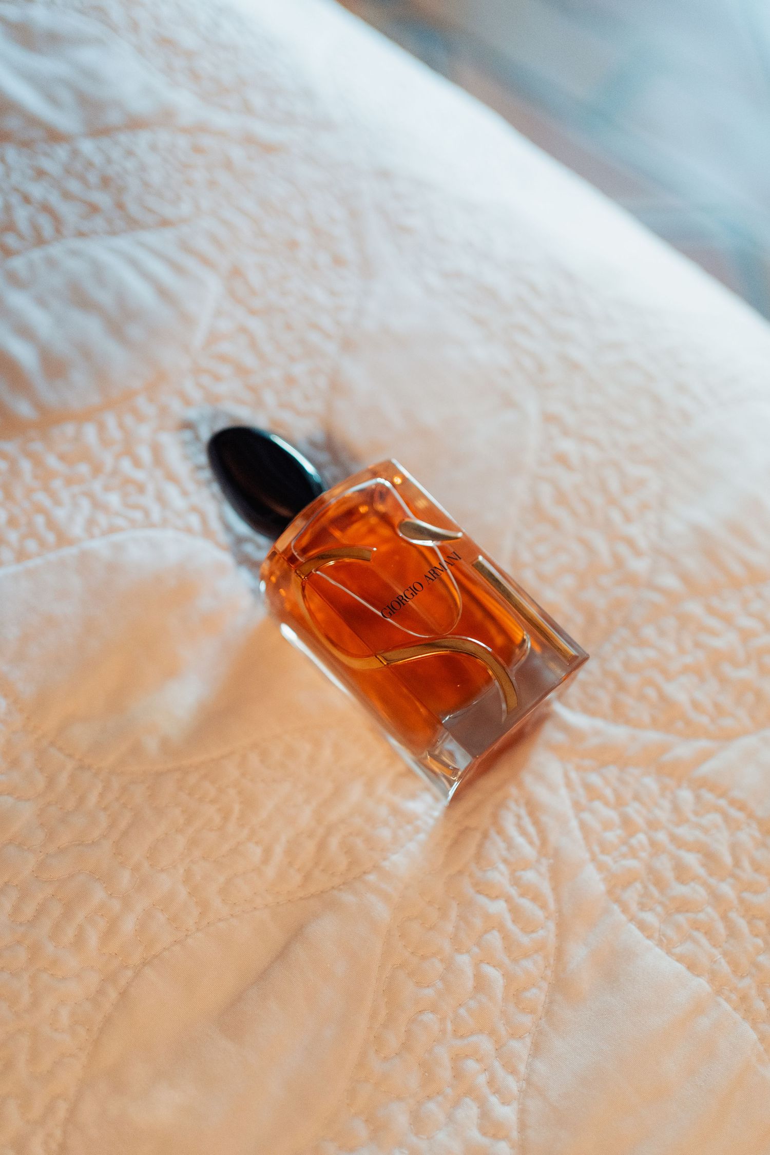 Amber perfume bottle with dropper cap and gold wave design on white quilted fabric surface.