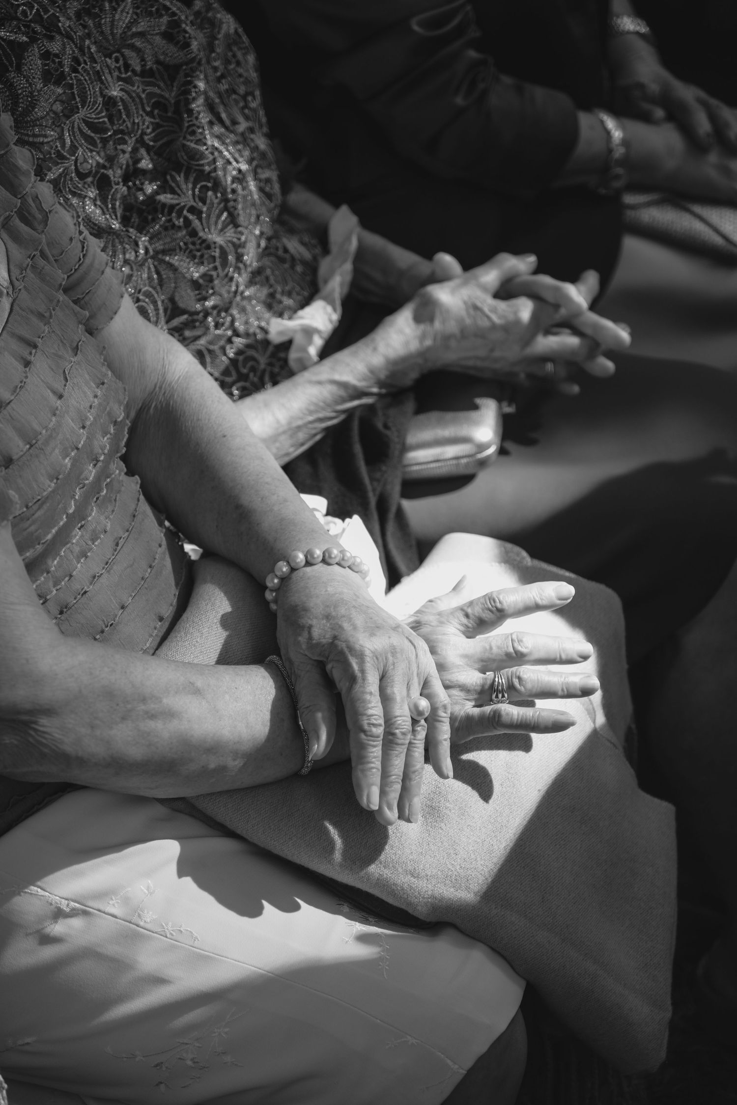 Black and white close-up of elderly women's hands with rings and bracelet clasped together at a wedding.