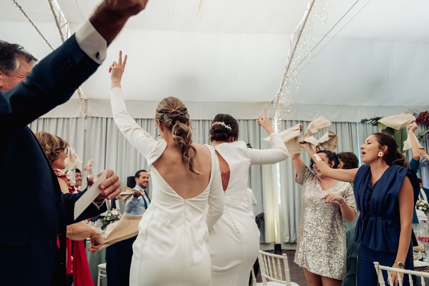Two brides in white wedding gowns walking through cheering guests inside a decorated reception marquee.
