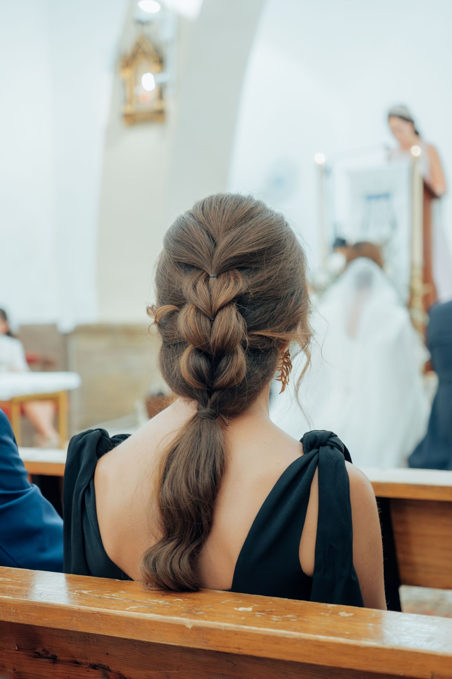 Woman with elegant braided ponytail hairstyle seated in church pew at wedding ceremony.