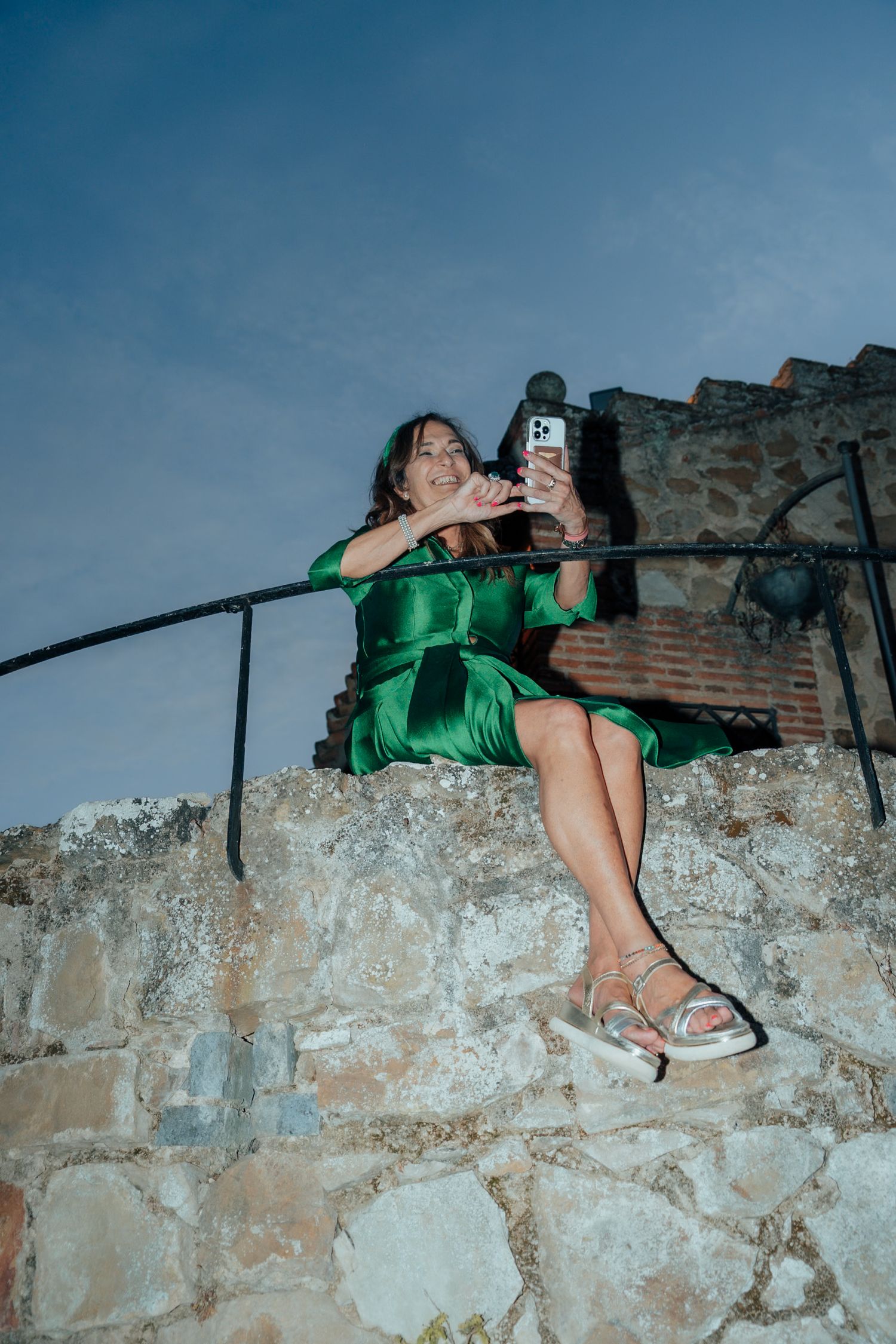 Woman in green satin dress sitting on stone wall taking selfie with smartphone at dusk.