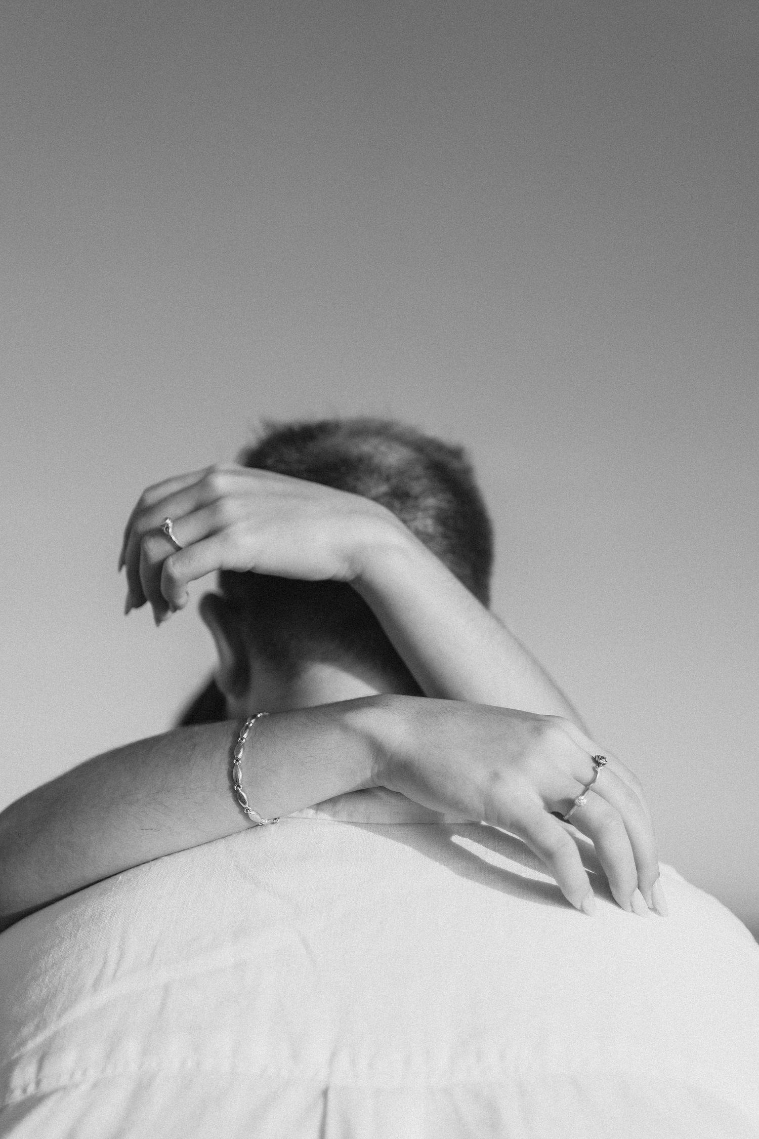 Black and white photo of a person being embraced from behind, showing hands with rings and a bracelet.