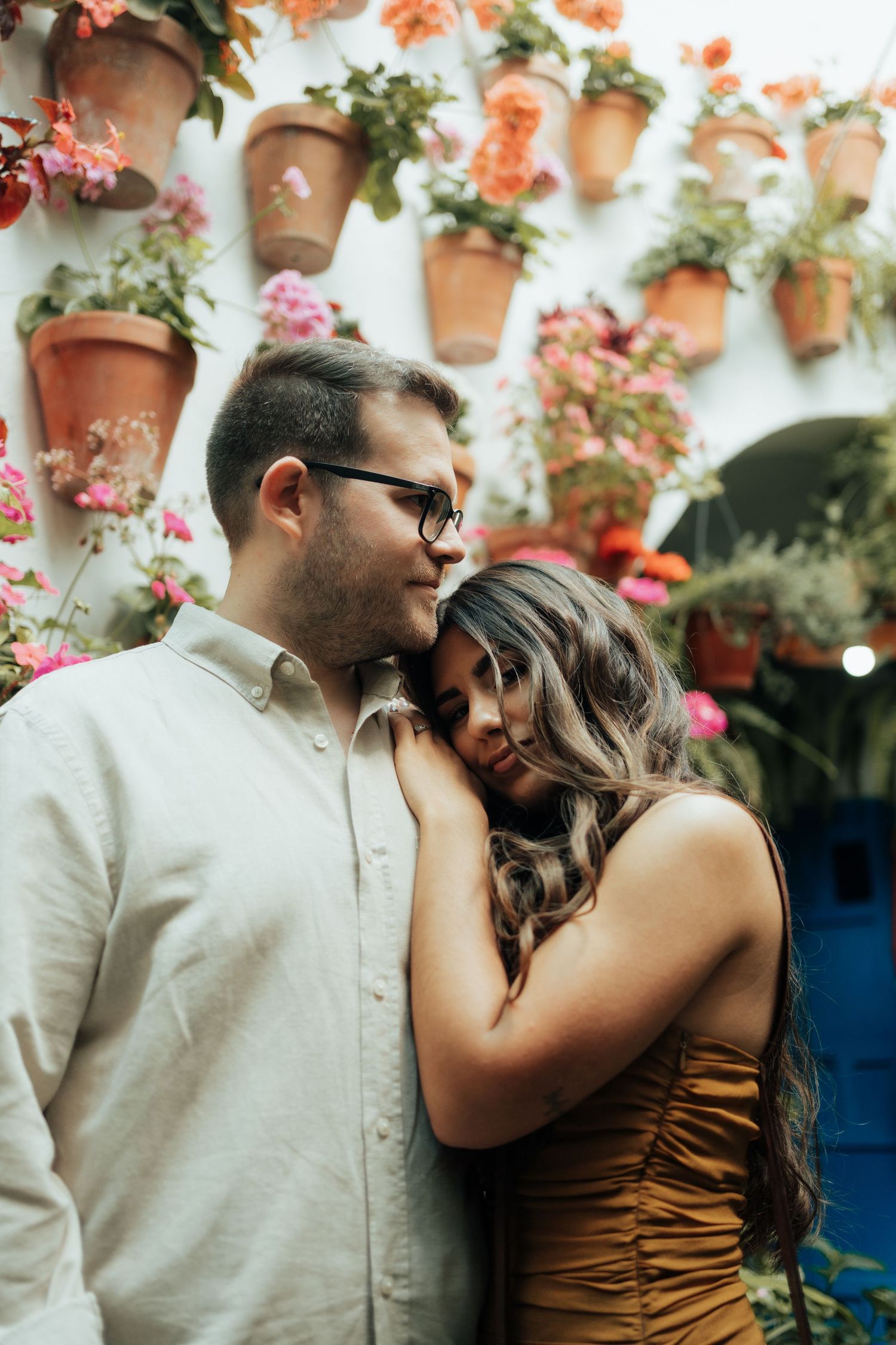 Romantic couple embracing in front of a white wall adorned with terracotta pots of colorful flowers.
