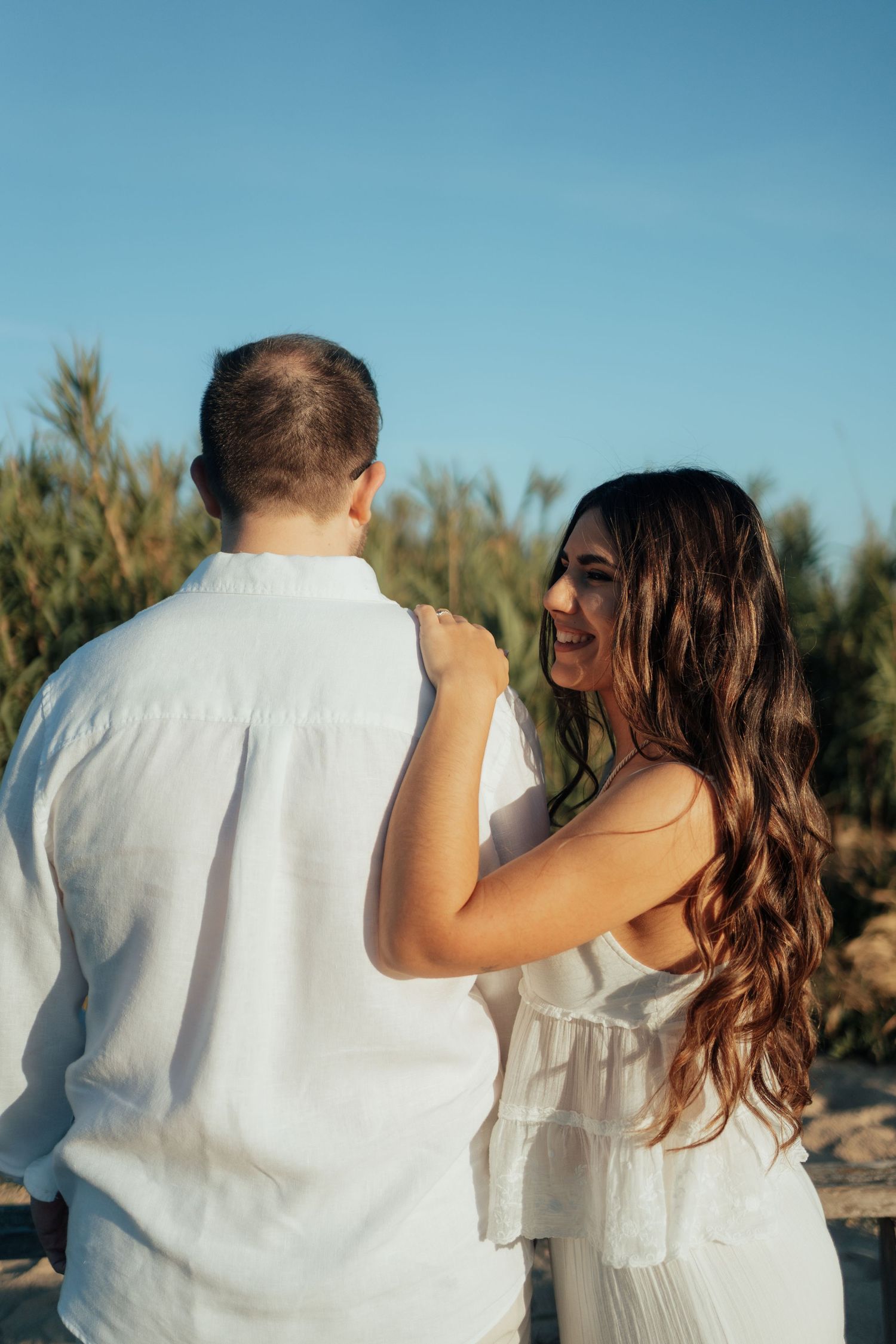 A smiling woman with long brown hair in a white dress rests her hand on a man's shoulder outdoors.