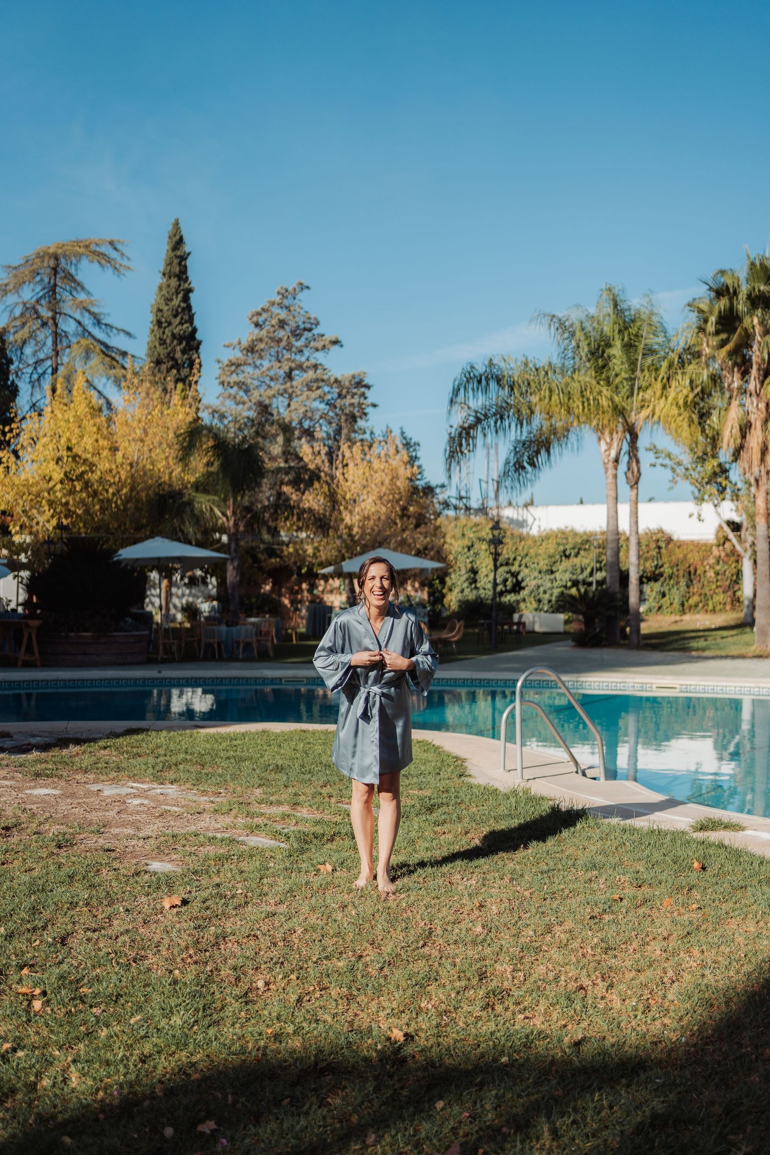 Joyful bride in blue satin robe standing barefoot by pool surrounded by palm trees.