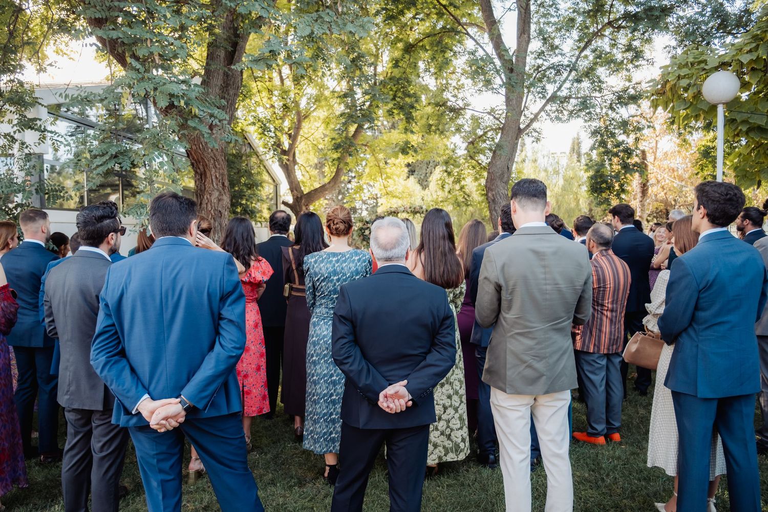 Wedding guests gathered outdoors under trees watching a garden ceremony from behind.
