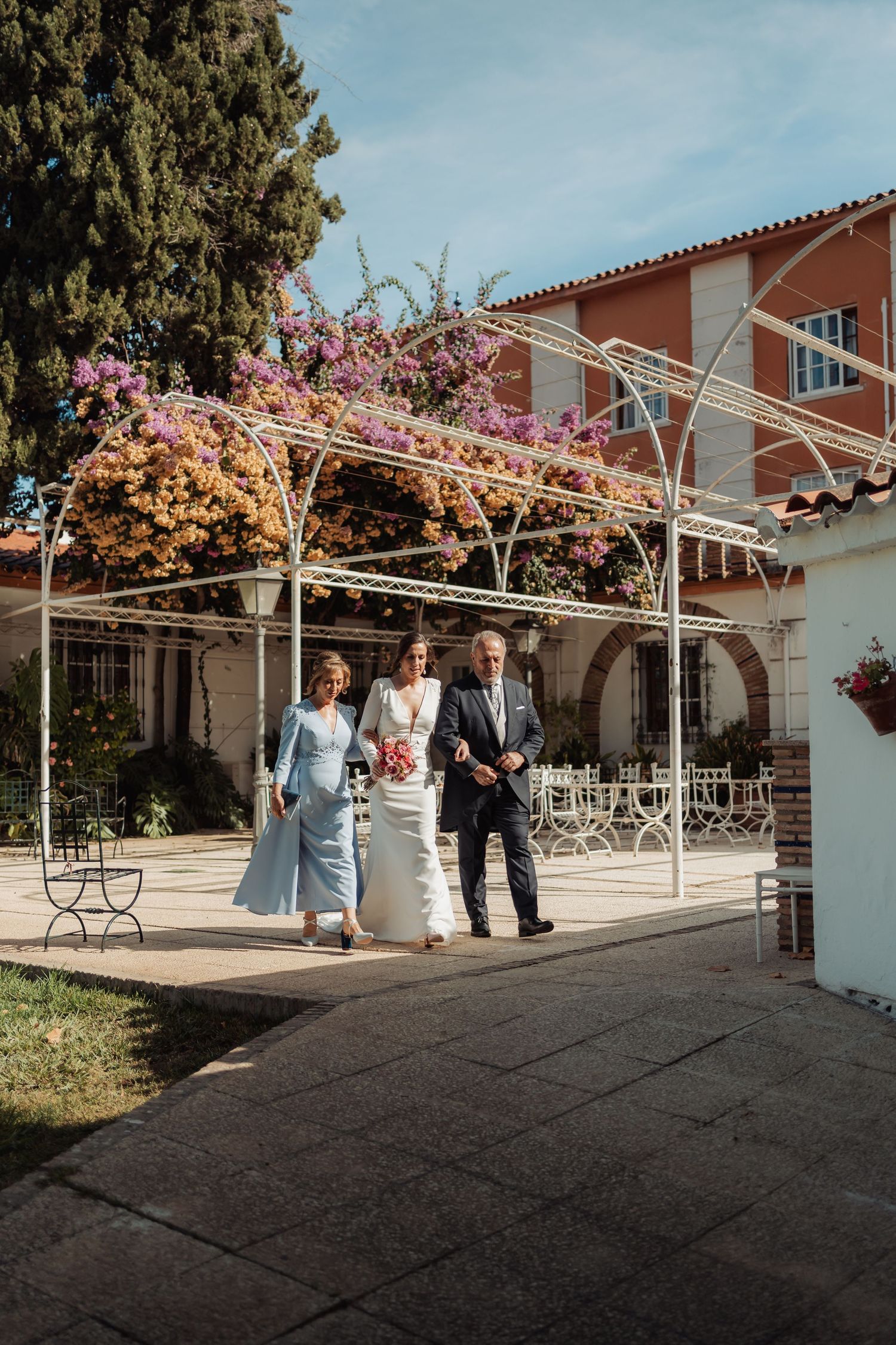 Bride escorted by parents walks under a white floral pergola toward the wedding ceremony.