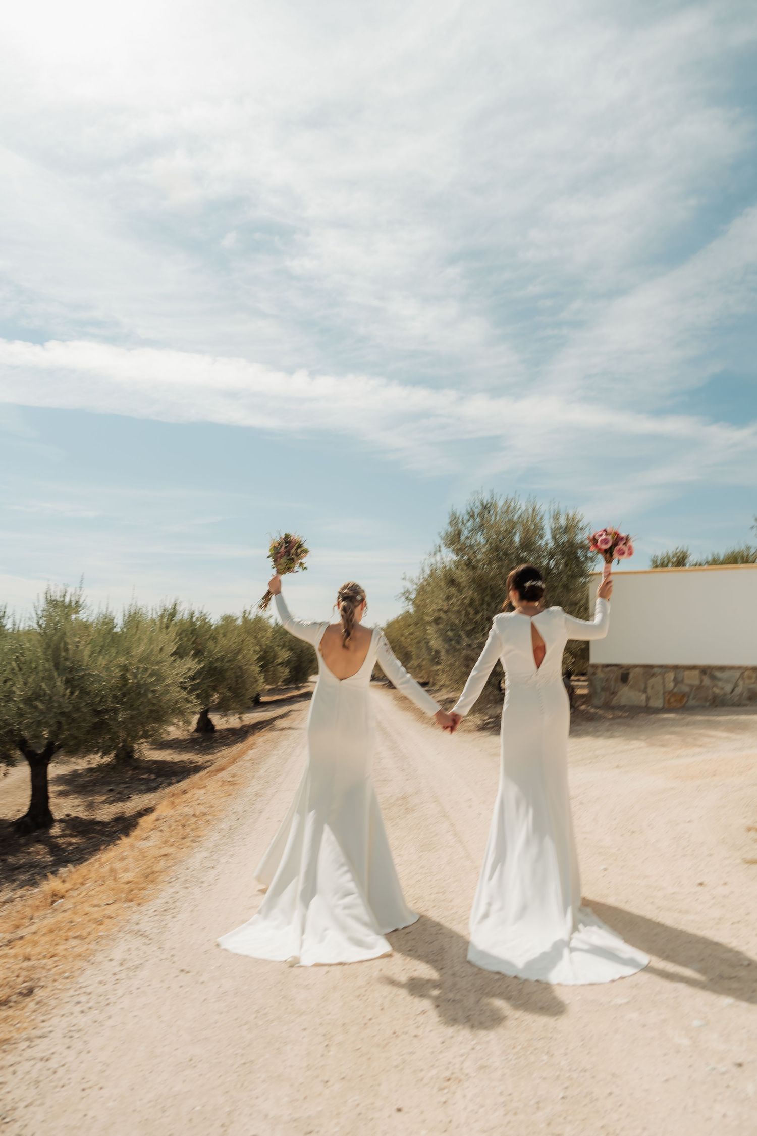 Two brides in elegant white dresses raising bouquets while holding hands in an olive orchard.