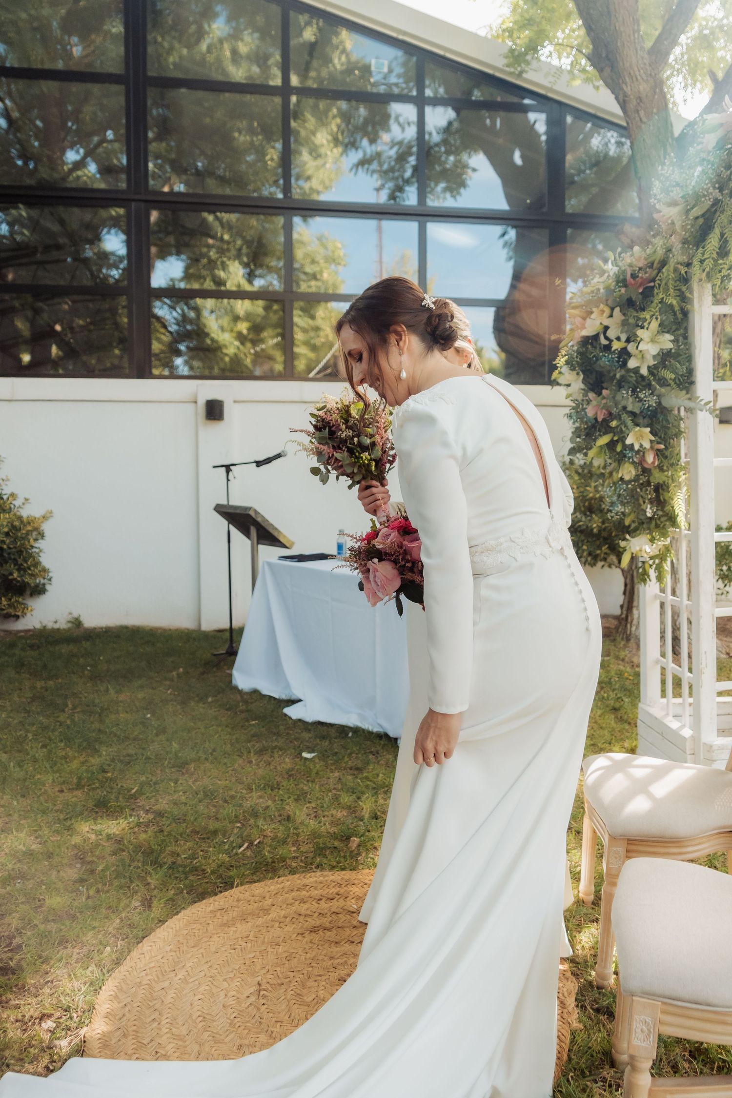 Elegant bride in long-sleeve white gown smells her colorful bouquet at outdoor wedding.
