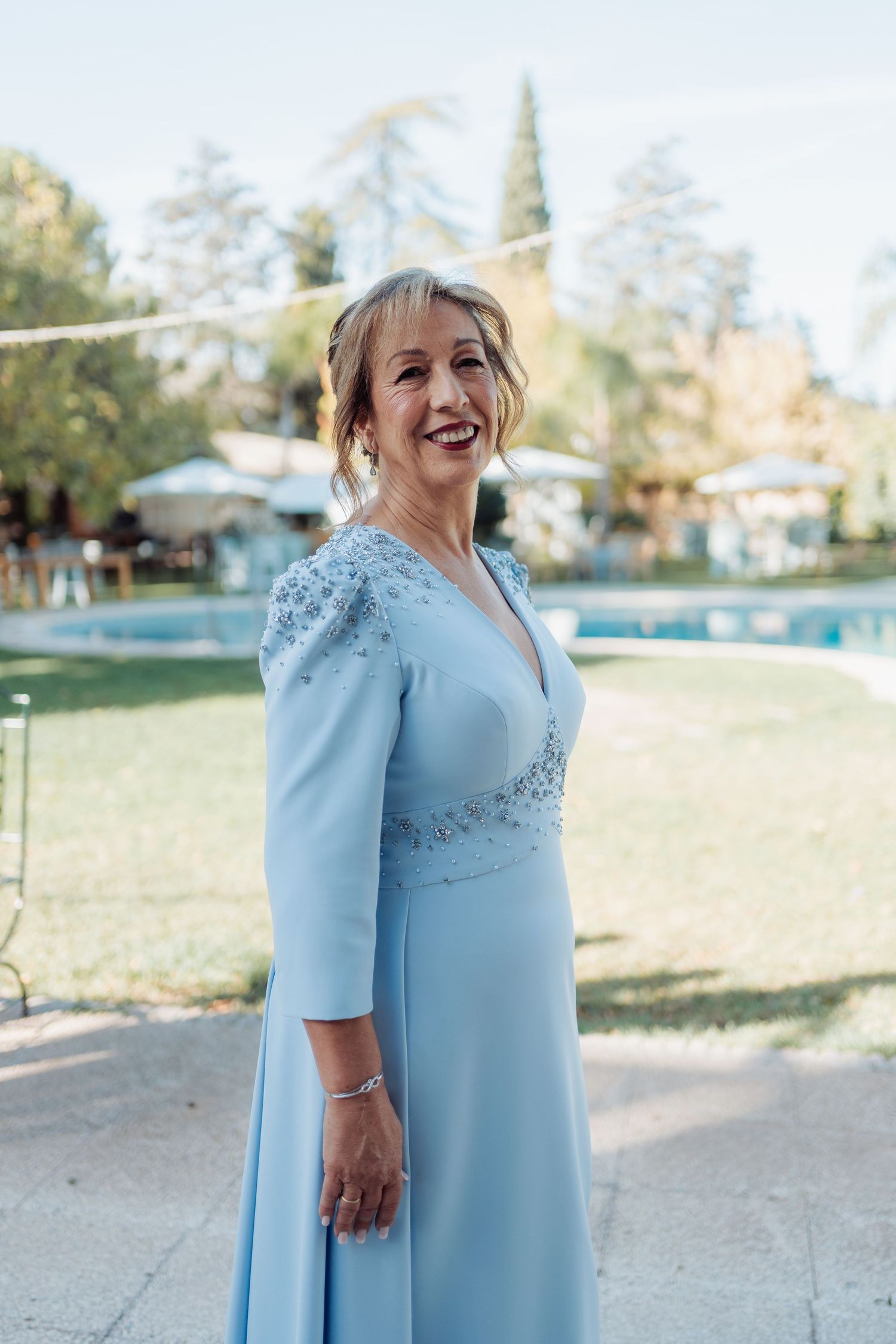 Elegant older woman in light blue beaded gown smiles outdoors near pool at wedding venue.