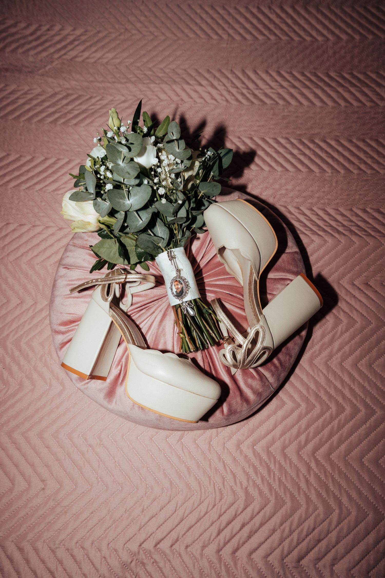 Bridal bouquet with eucalyptus and cream platform heels arranged on a pink velvet pillow on quilted bedding.