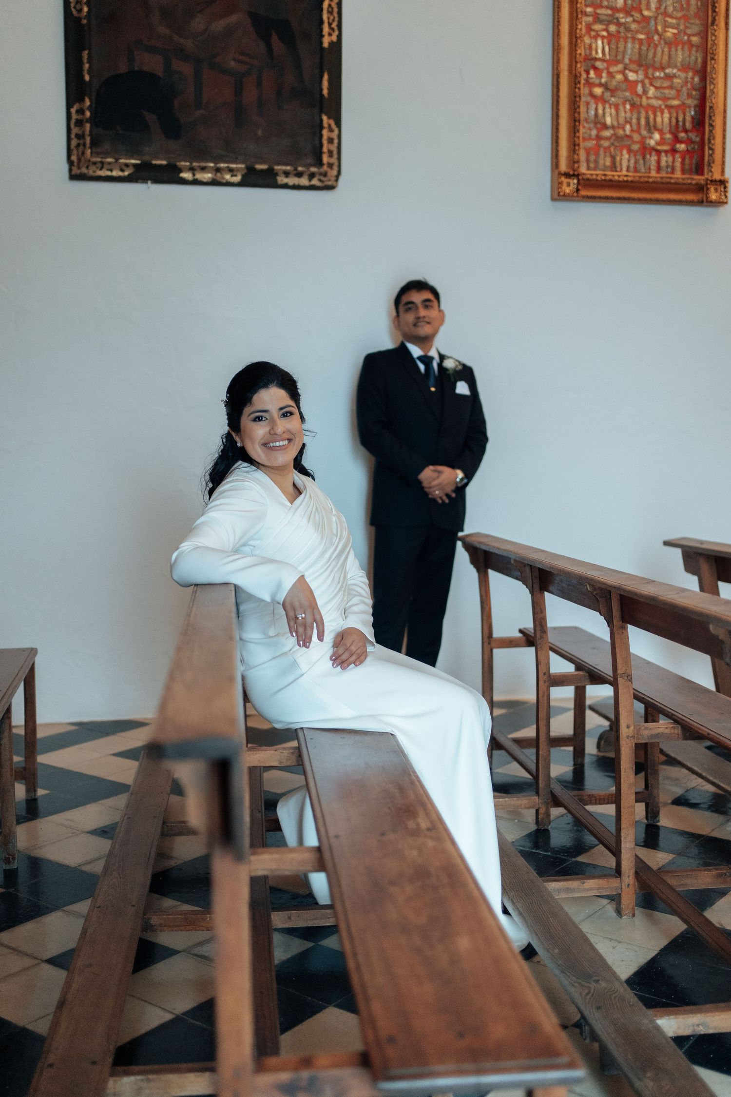 Smiling bride in white gown sitting on church pew with groom standing behind her.