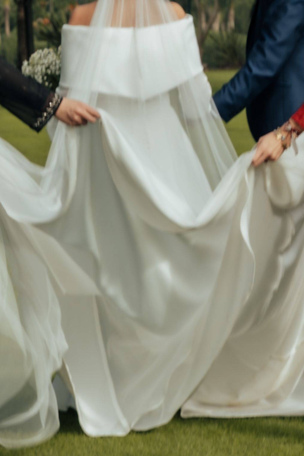 Close-up of wedding party holding the bride's flowing white dress and veil train on green grass.