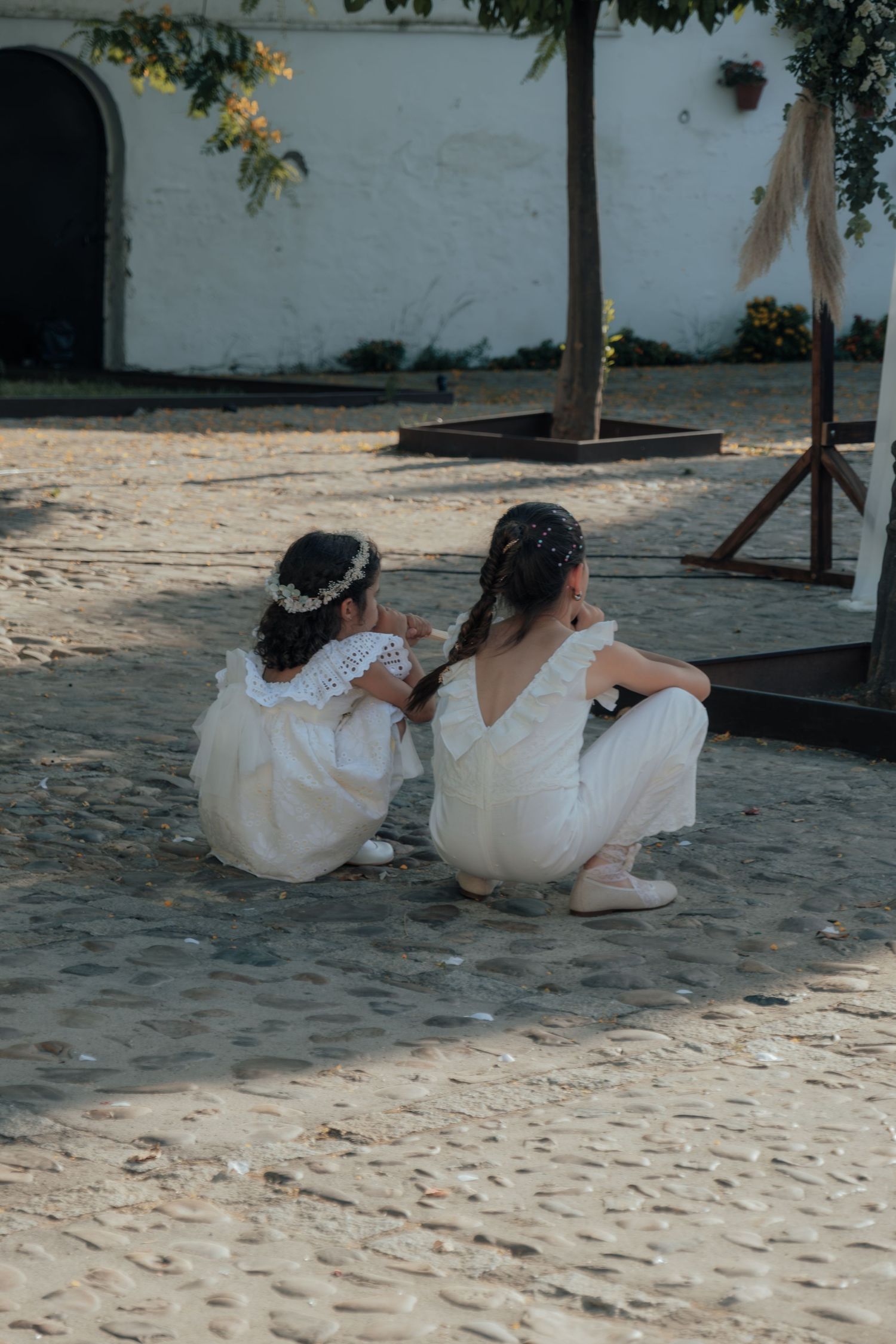Two young flower girls in white dresses crouch together on cobblestone at an outdoor wedding ceremony.