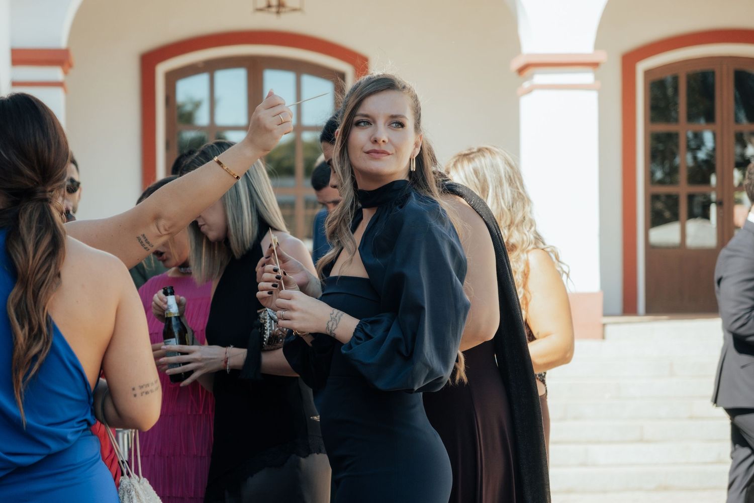 Woman in black dress with sheer sleeves socializing at outdoor wedding reception.