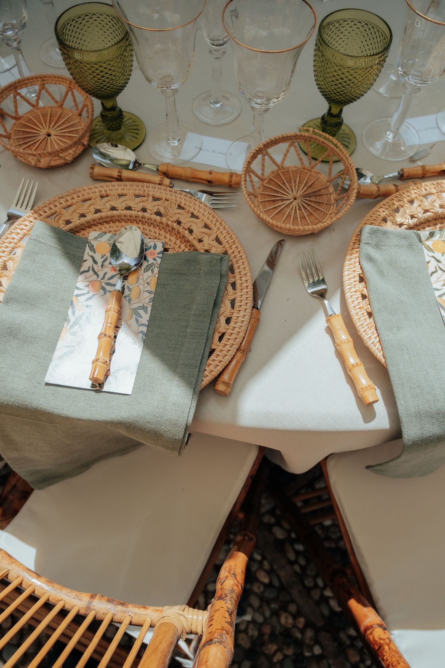 Top view of elegant wedding table setting with wicker chargers, green goblets, bamboo cutlery, and sage napkins.