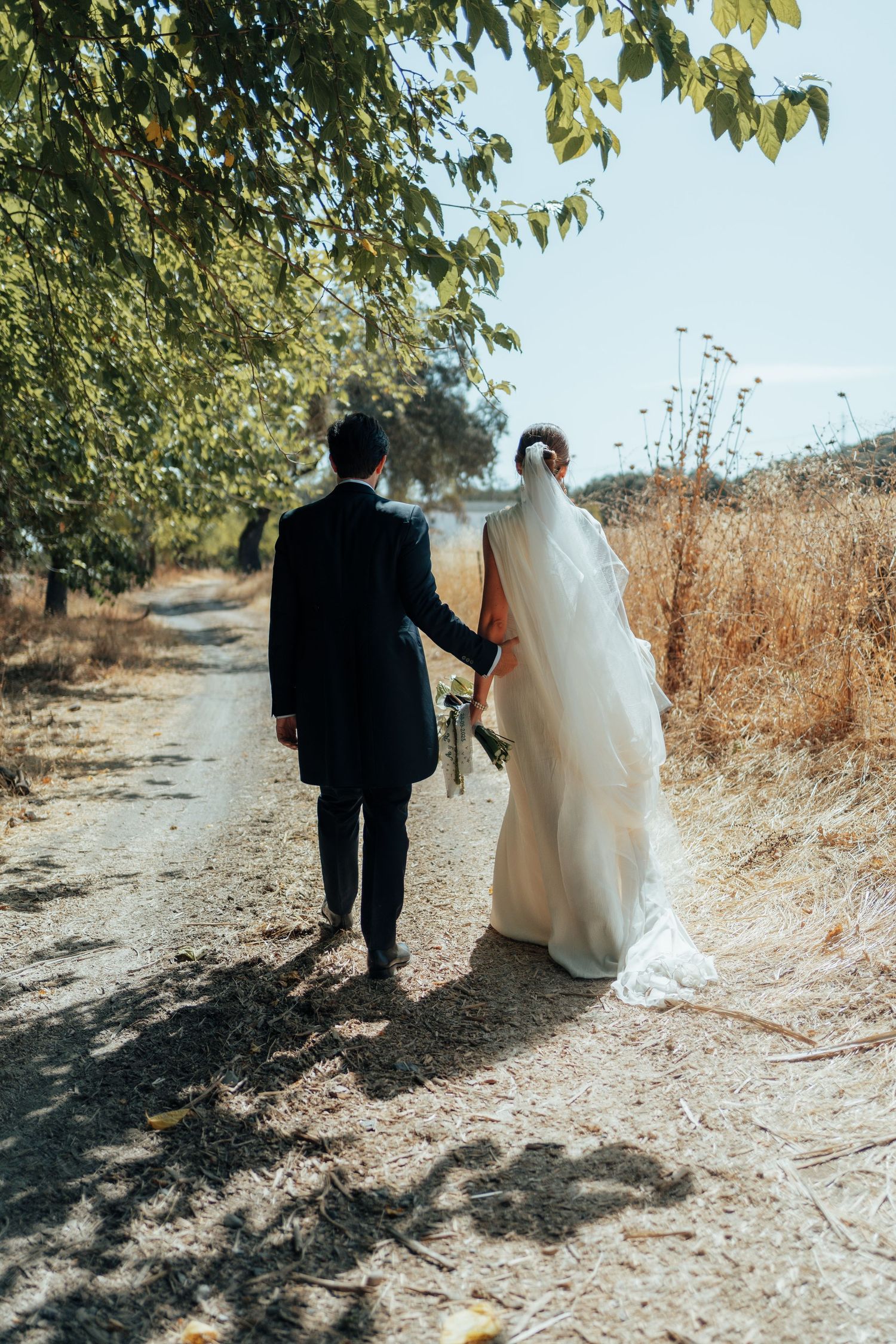 Wedding couple walking away on a sunny dirt path, bride in white gown with veil and groom in dark suit.